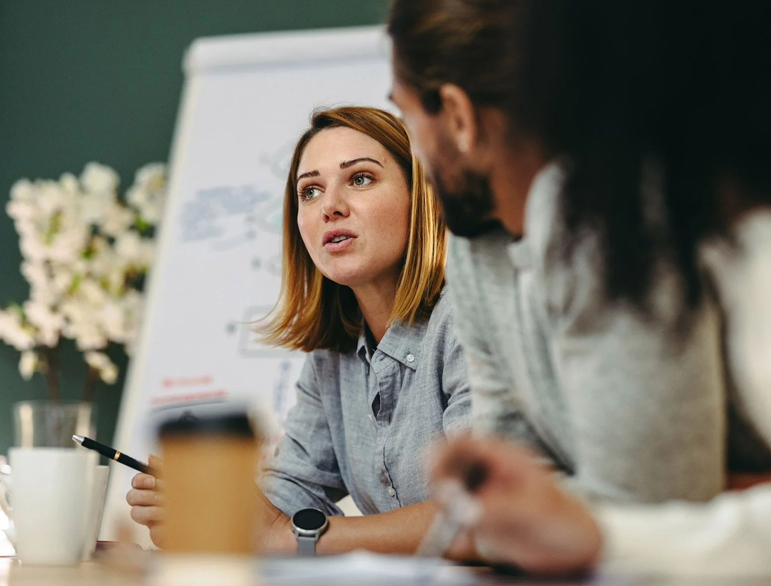 Two people in a meeting or discussion about human resources and health and safety issues, one woman with red hair speaking and holding a pen, another man with dark hair listening, in a conference room with a whiteboard and flowers in the background.