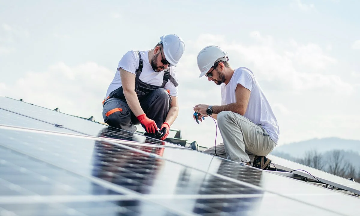Two workers wearing hard hats and safety glasses installing or inspecting solar panels on a rooftop.