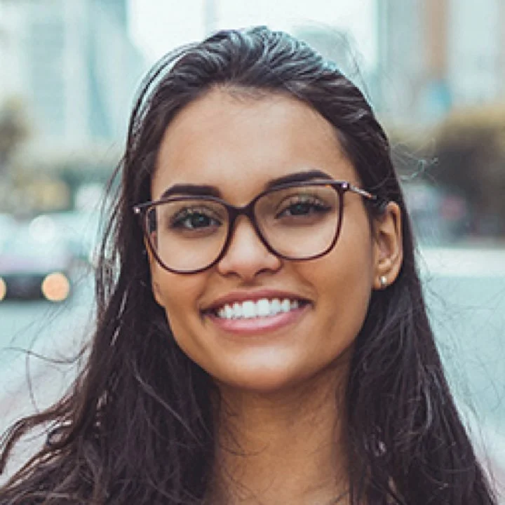 A young woman with long dark hair and glasses smiling outdoors in an urban setting.