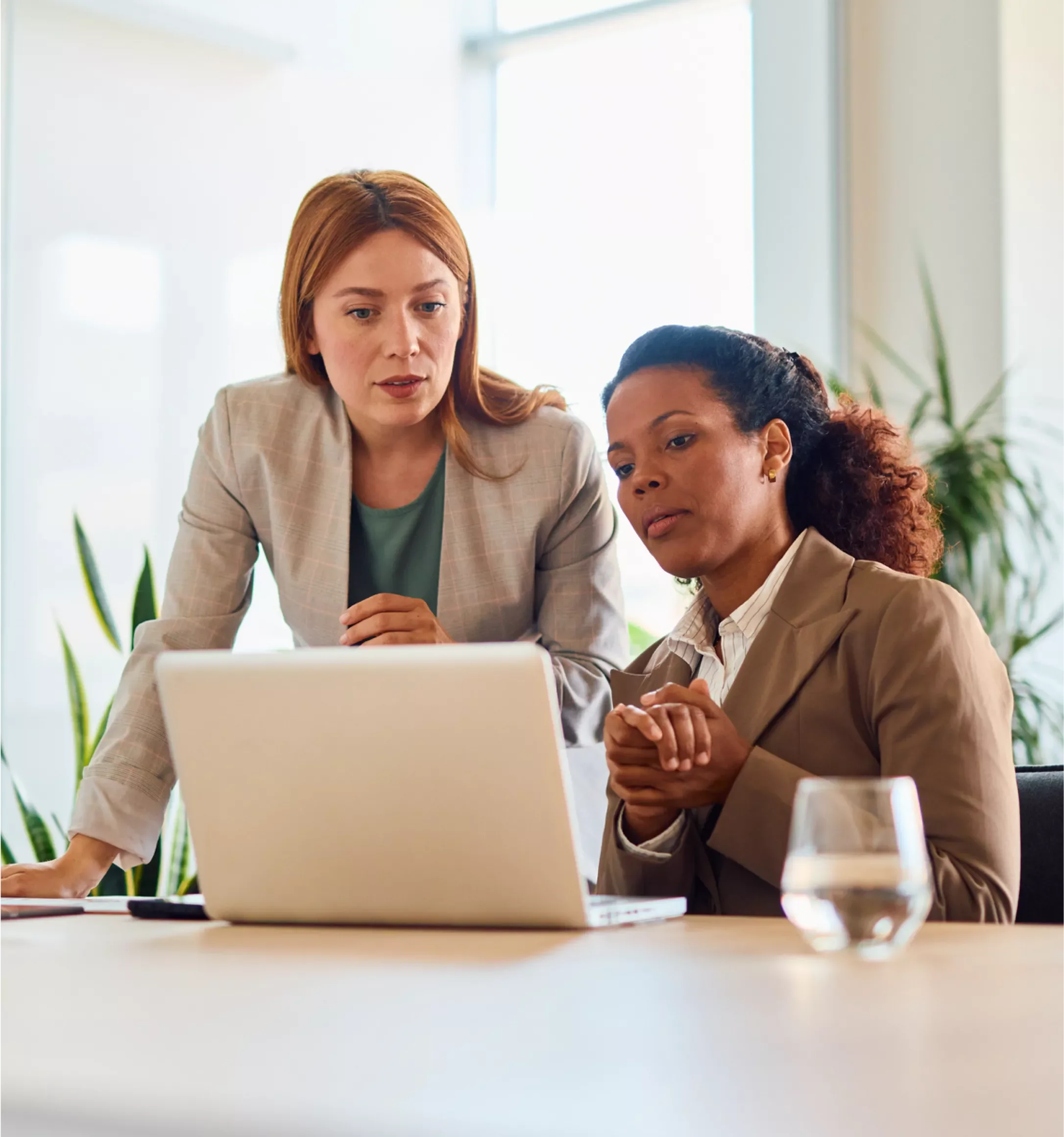 Two women, one with red hair and one with curly dark hair, work together on a laptop in a bright office space.
