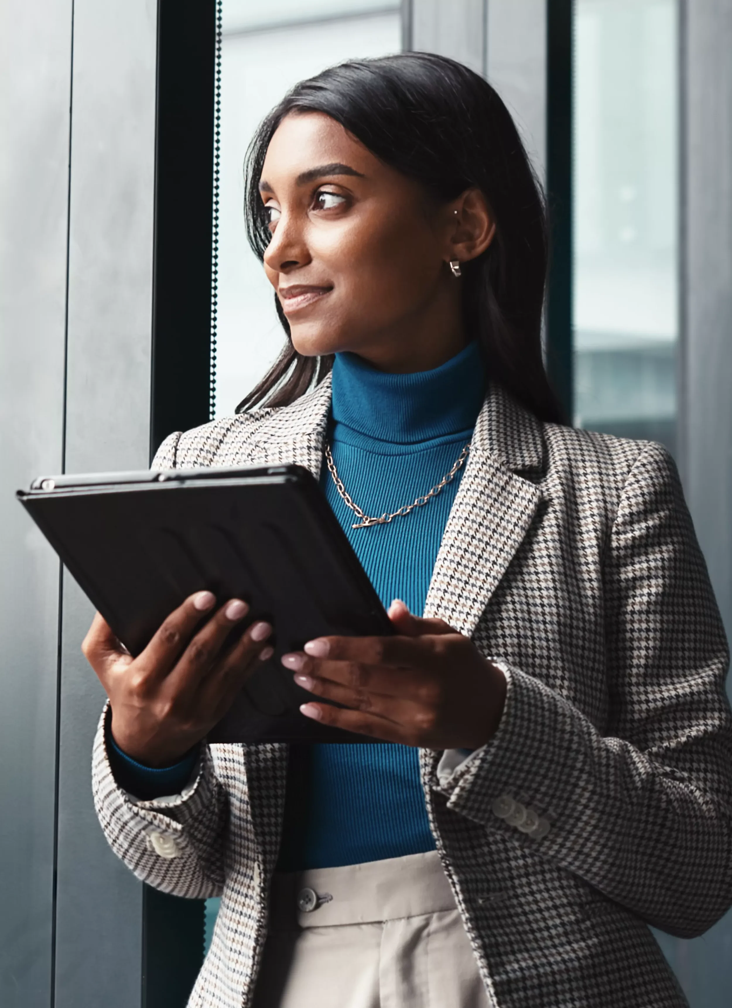 A professional woman with dark hair, wearing a checkered blazer and blue turtleneck, holding a tablet while standing in an elevator.