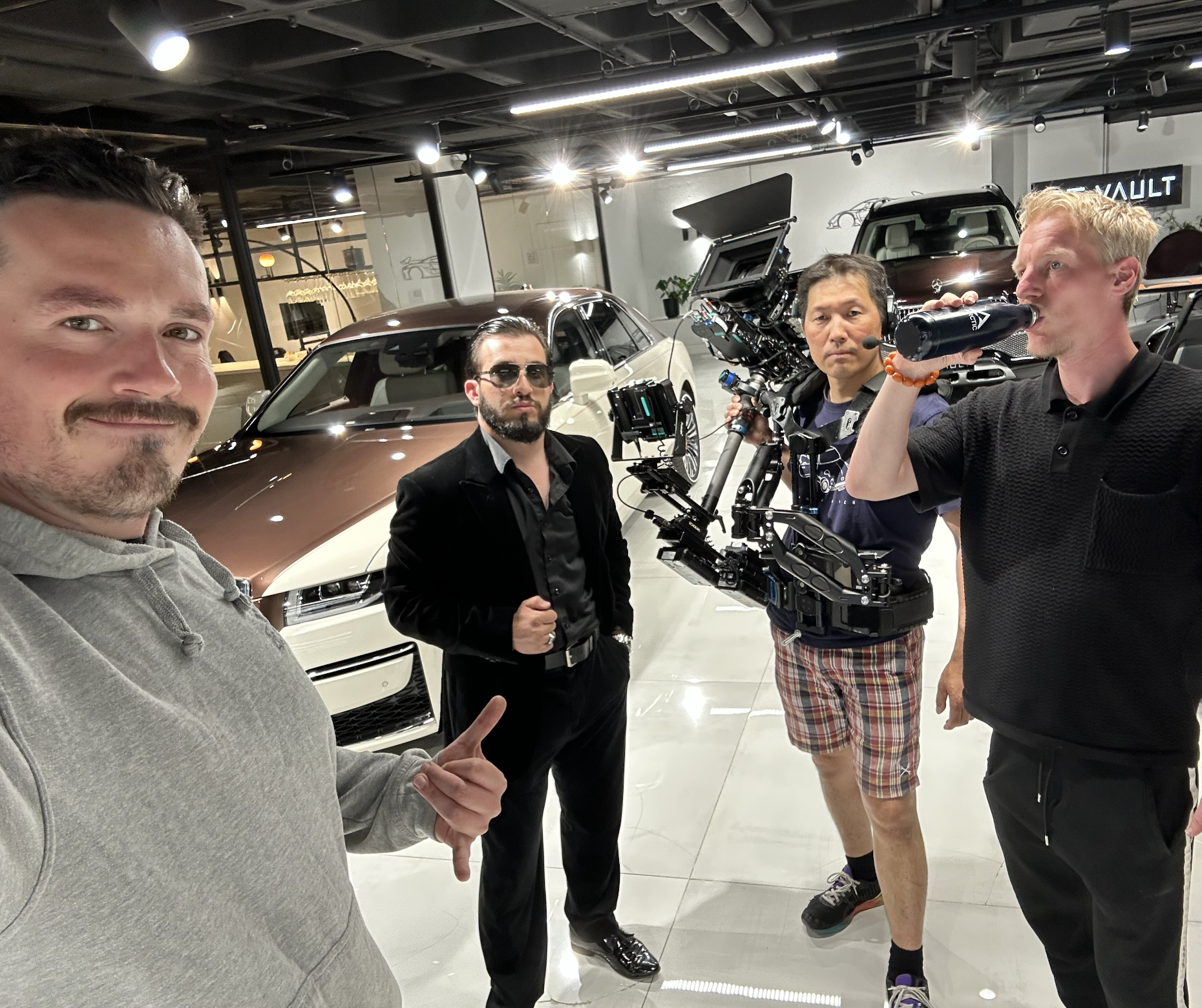 Four men posing inside a car dealership with a brown and white car behind them. One person is taking a selfie with the group. The other three men are holding filming equipment or a drink, with one wearing sunglasses and a black suit, another in black polo with curly hair drinking from a bottle, and the third with a camera rig.