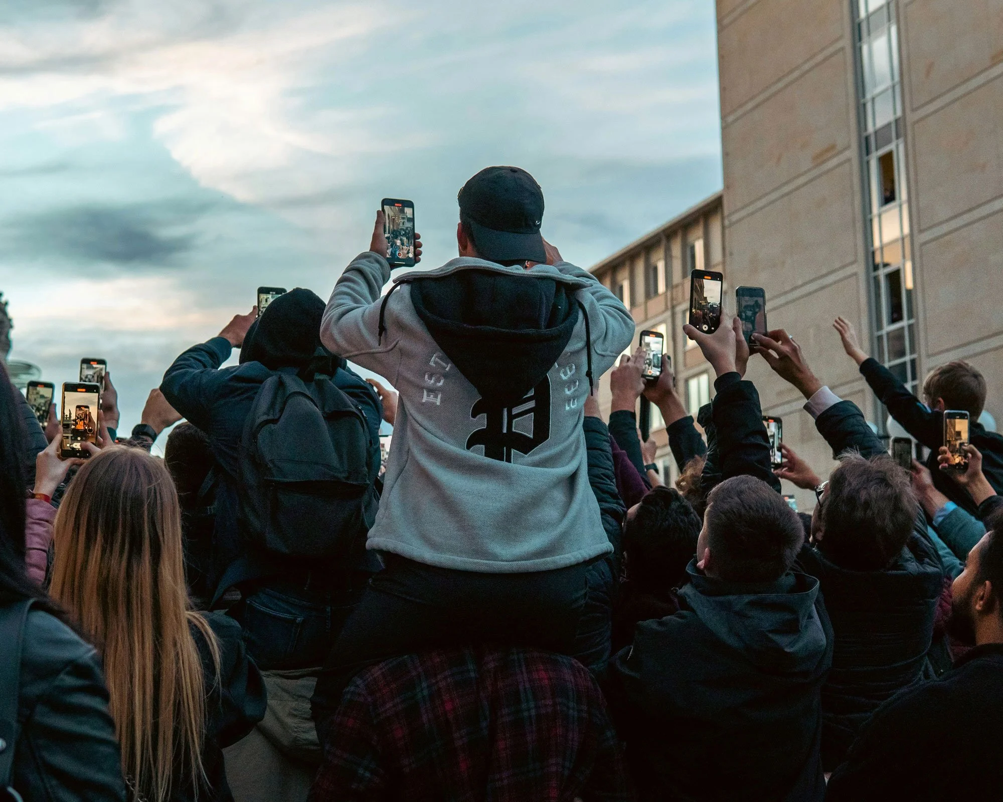 Crowd of people outdoors taking photos or videos with smartphones, some standing, some sitting, near a building under a cloudy sky.