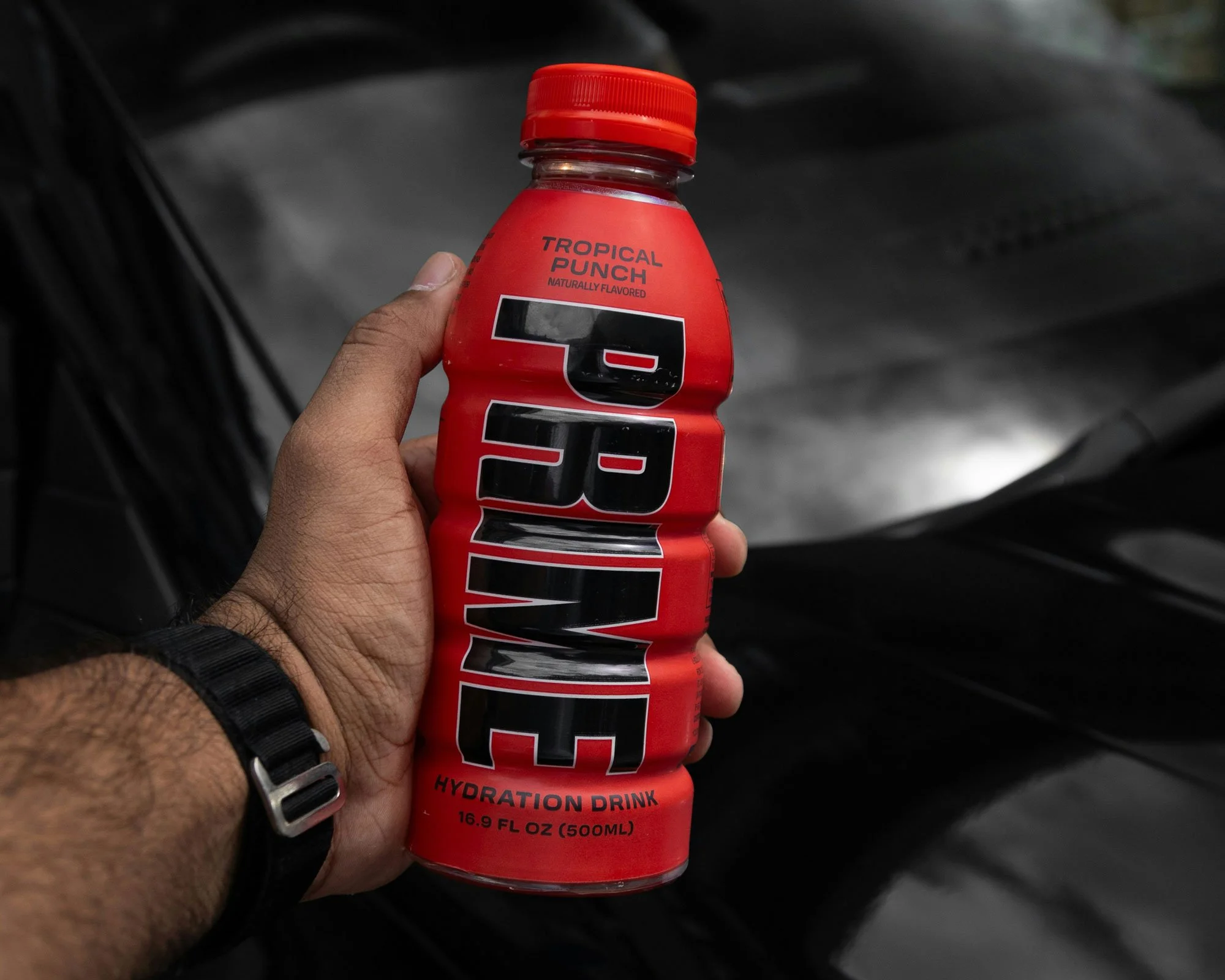 A hand holding a red bottle of 5-hour Energy Tropical Punch hydration drink. The bottle has a black, white, and red label and a red cap, with a dark background.