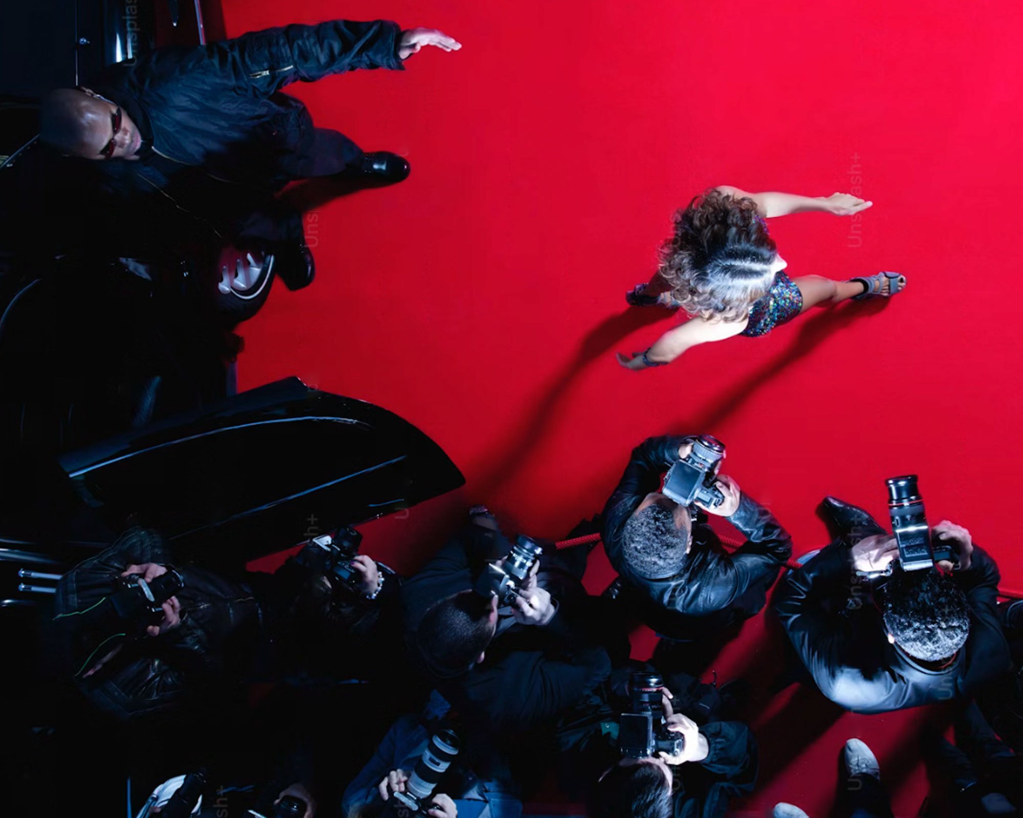 A woman with curly hair in a colorful dress poses confidently on a red carpet, surrounded by multiple photographers capturing her photo.