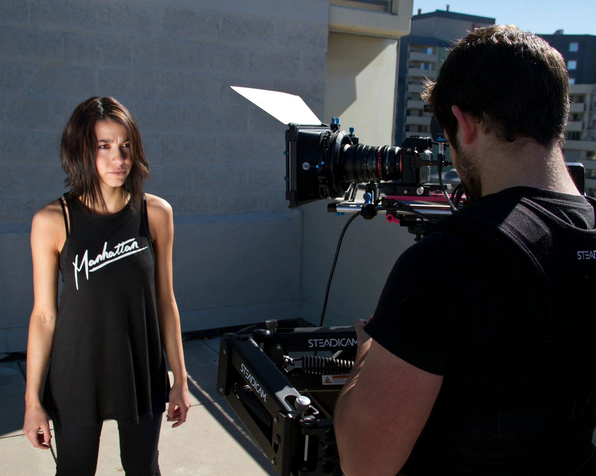 A woman with shoulder-length brown hair wearing a black tank top with 'Manhattan' written on it, standing outdoors in sunlight, being filmed by a man operating a professional camera mounted on a Steadicam. The background shows a cityscape with buildings.