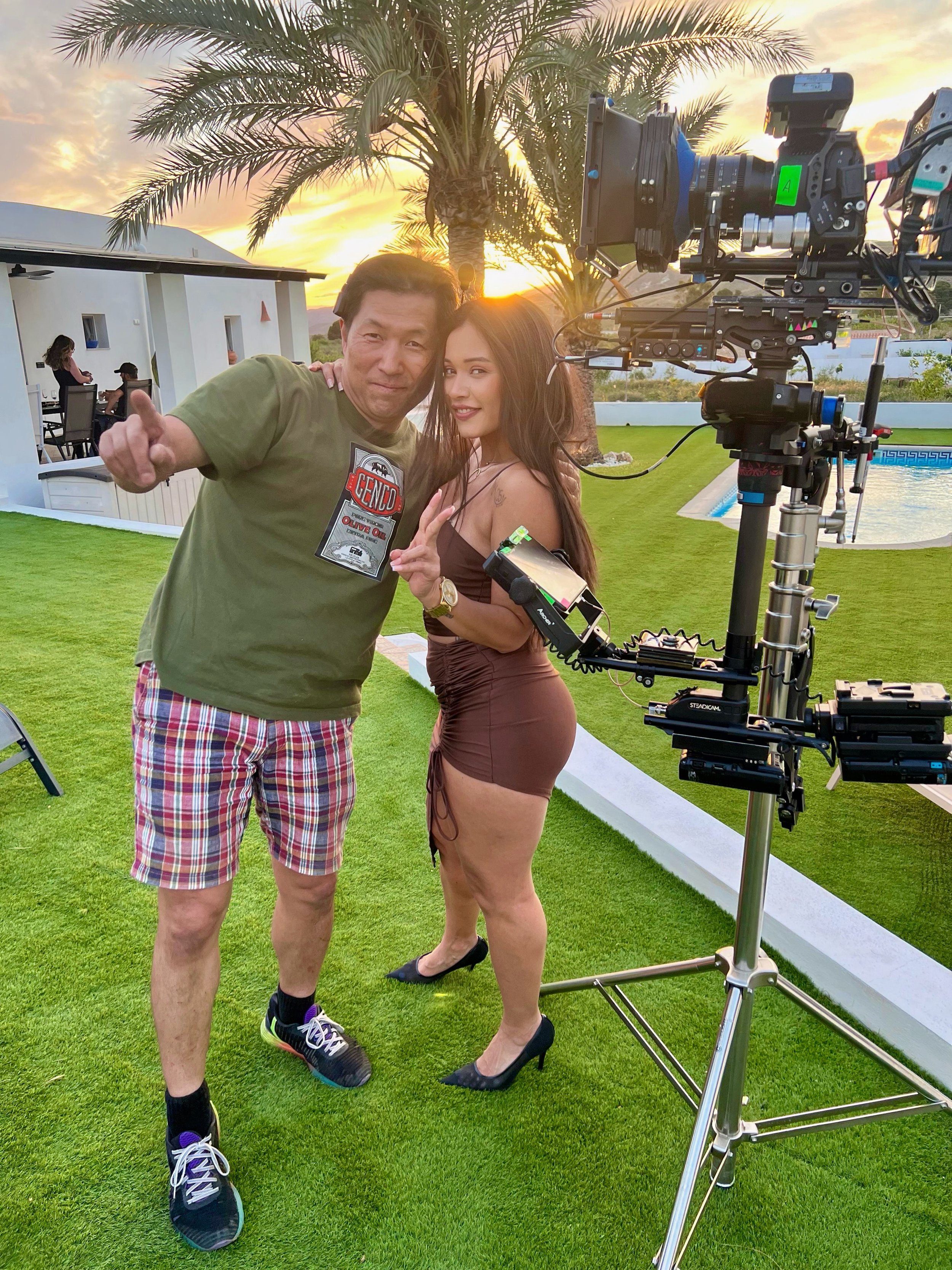 A man and woman standing on a lawn with a camera setup, sunset, palm trees, swimming pool, and a white building in the background.