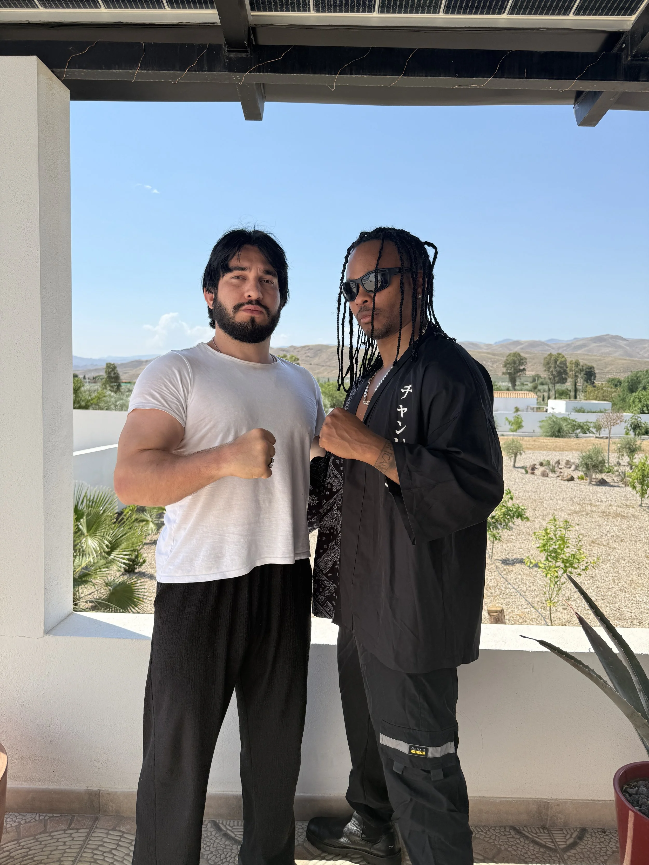 Two men standing outside on a balcony, posing with their fists raised, with a landscape of hills and trees in the background, under a clear blue sky.