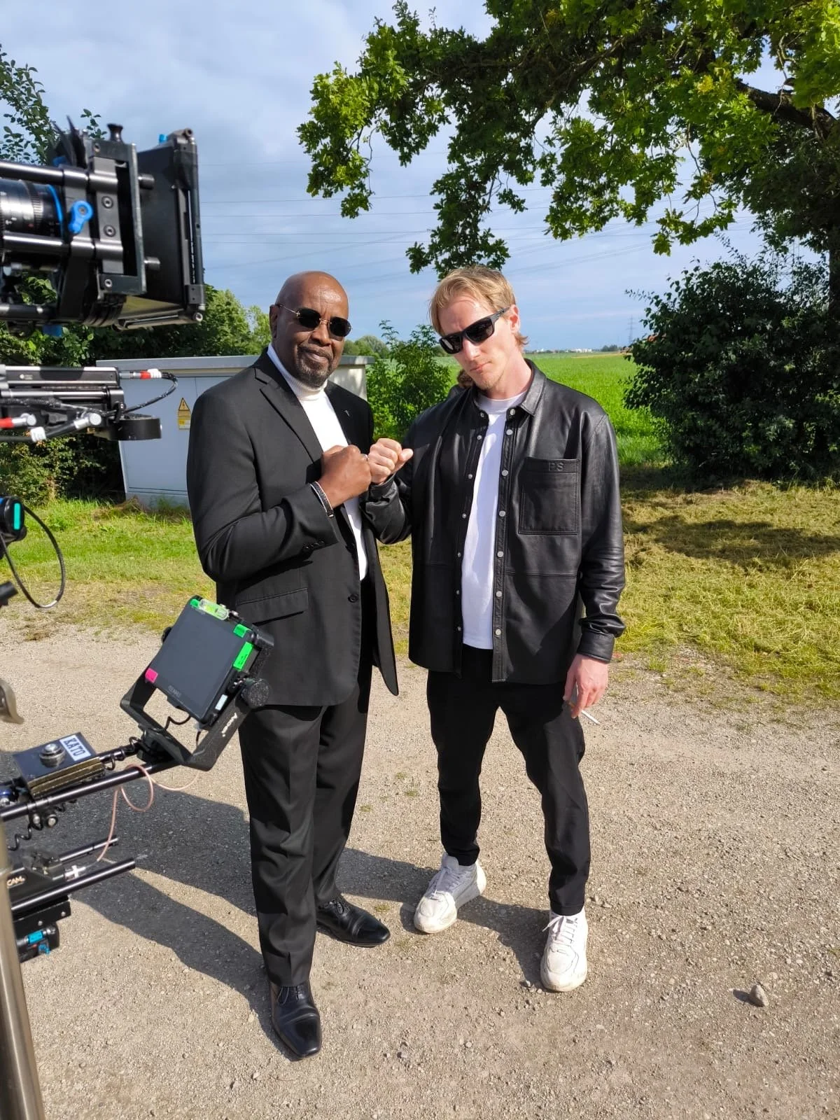 Two men in sunglasses outdoors, fist bumping. One in a black suit and white shirt, the other in a black leather jacket and white sneakers. Film set equipment visible on the left, green trees and field in the background.