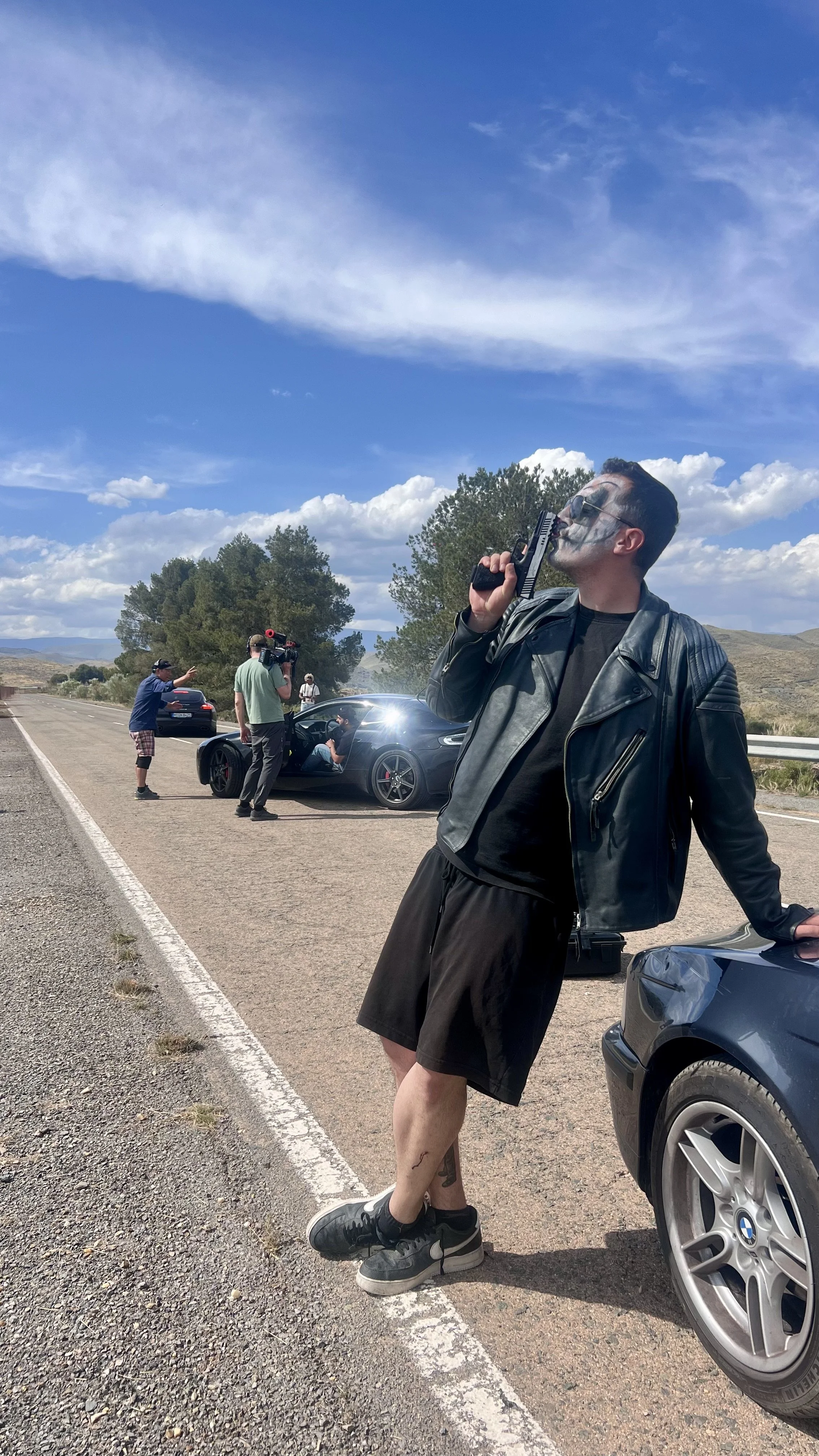 A man in a black leather jacket and shorts leaning on a car holding a gun to his mouth, with a crew filming a scene involving a sports car and actors in the background on a rural road.
