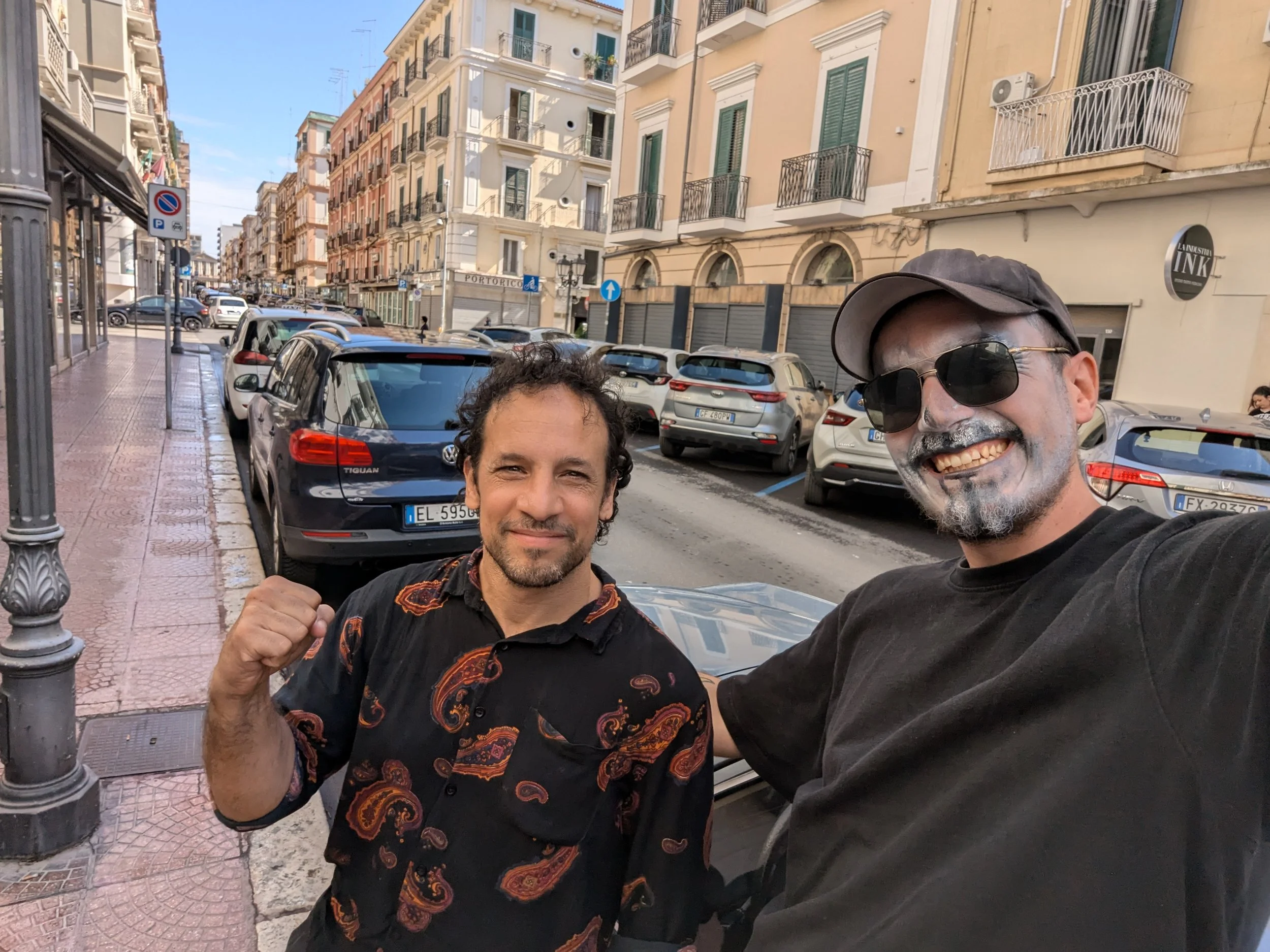 Two men taking a selfie on a city street with parked cars and multi-story buildings in the background. One man is wearing a black shirt with a colorful pattern, and the other is wearing sunglasses, a cap, and a black t-shirt. One is making a fist gesture.