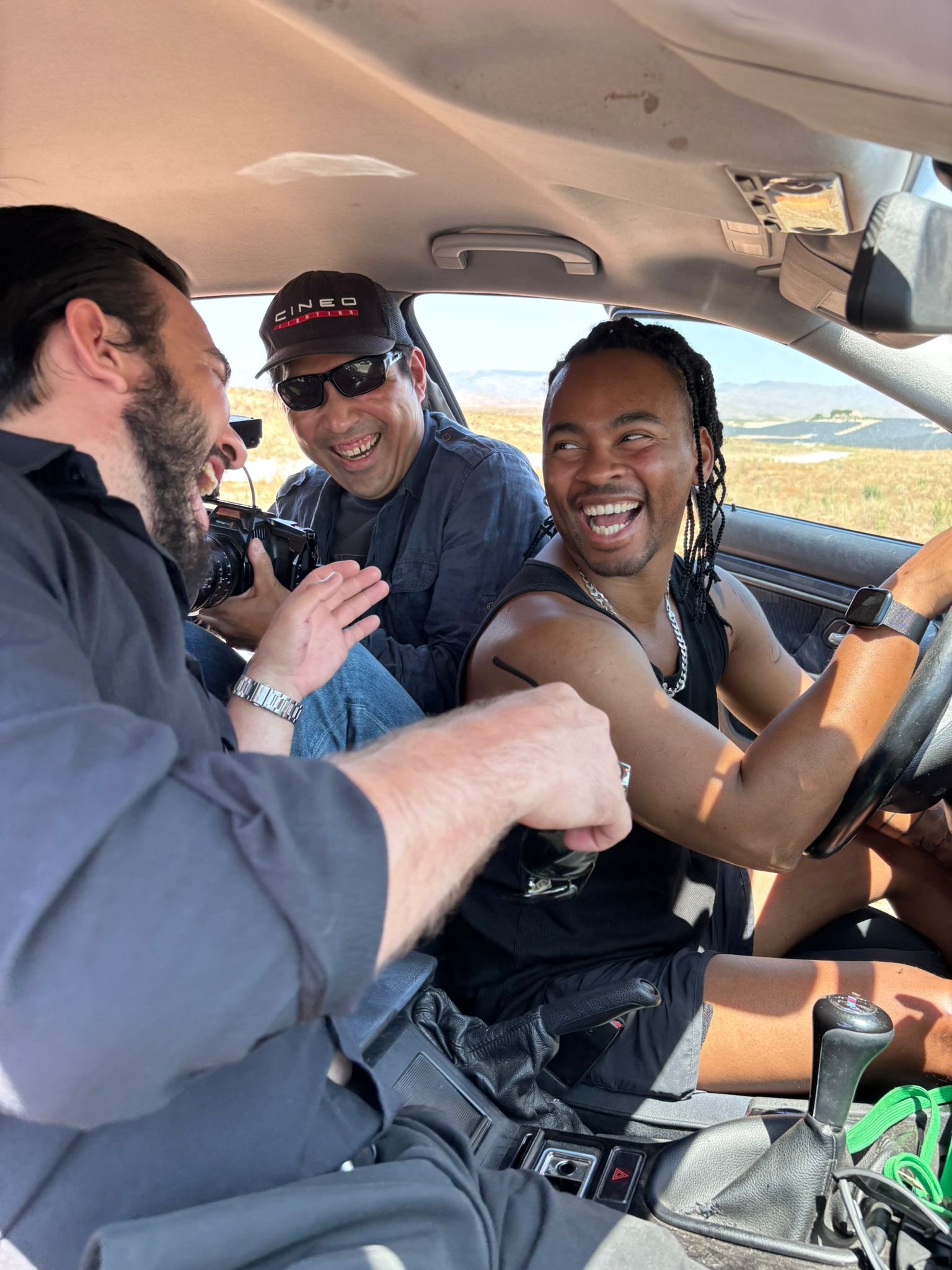 Three men inside a vehicle laughing and smiling, with one man in the driver's seat and the other two in the back.