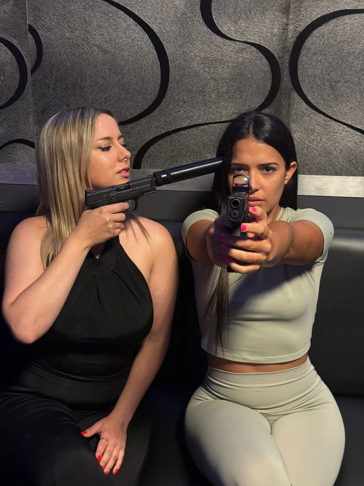 Two women sitting on a black couch with a textured wall in the background, one woman with blonde hair has a gun to the other woman's temple, who is pointing a gun directly at the camera.