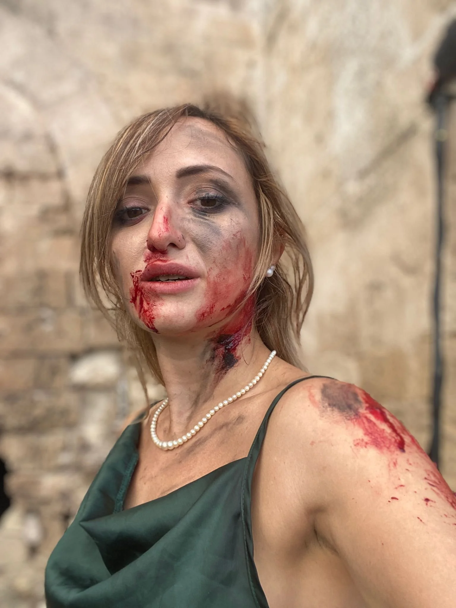 A woman with makeup and fake blood on her face, shoulder, and neck, wearing a pearl necklace and a dark green dress, standing against a stone wall.