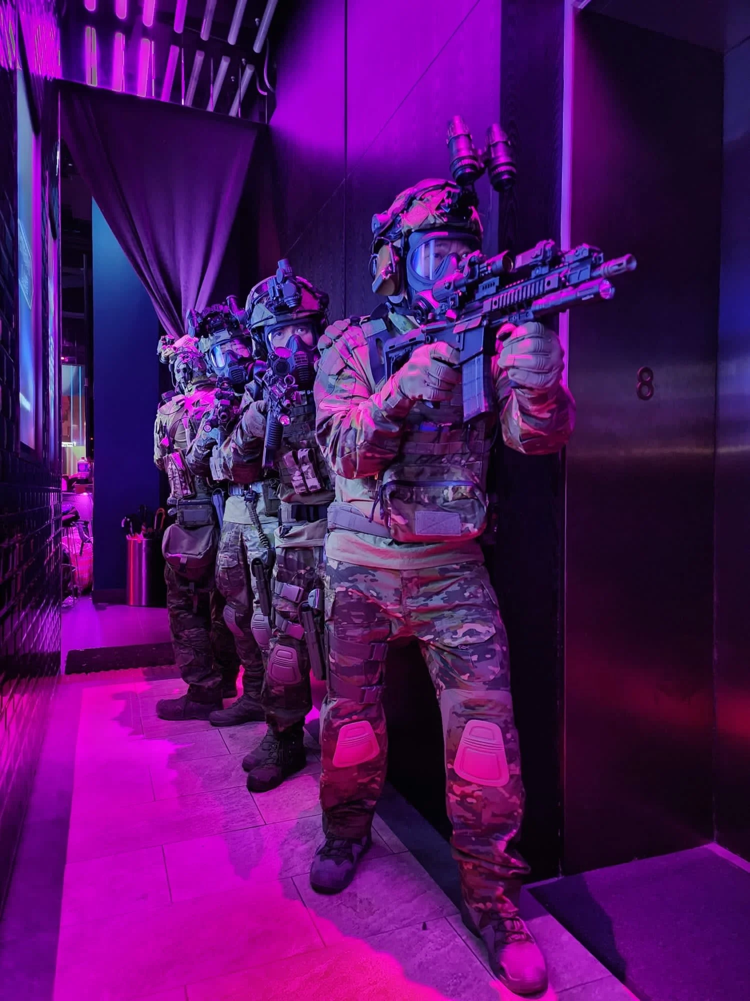 Three individuals dressed in military tactical gear, including camouflage uniforms and helmets with night vision goggles, are lined up inside a dimly-lit room with purple lighting, holding rifles and ready for action.