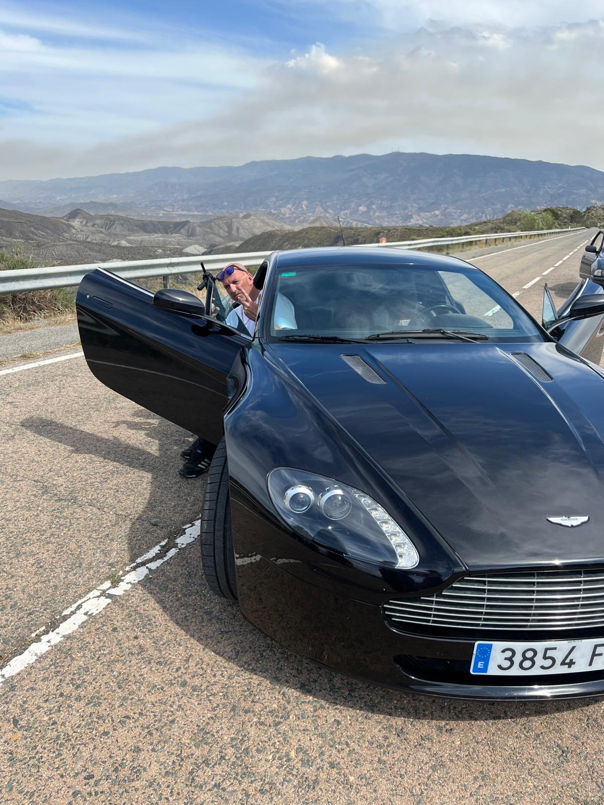 A man with sunglasses leaning out of a black Aston Martin sports car on the side of a scenic mountain road, with mountainous terrain and a cloudy sky in the background.