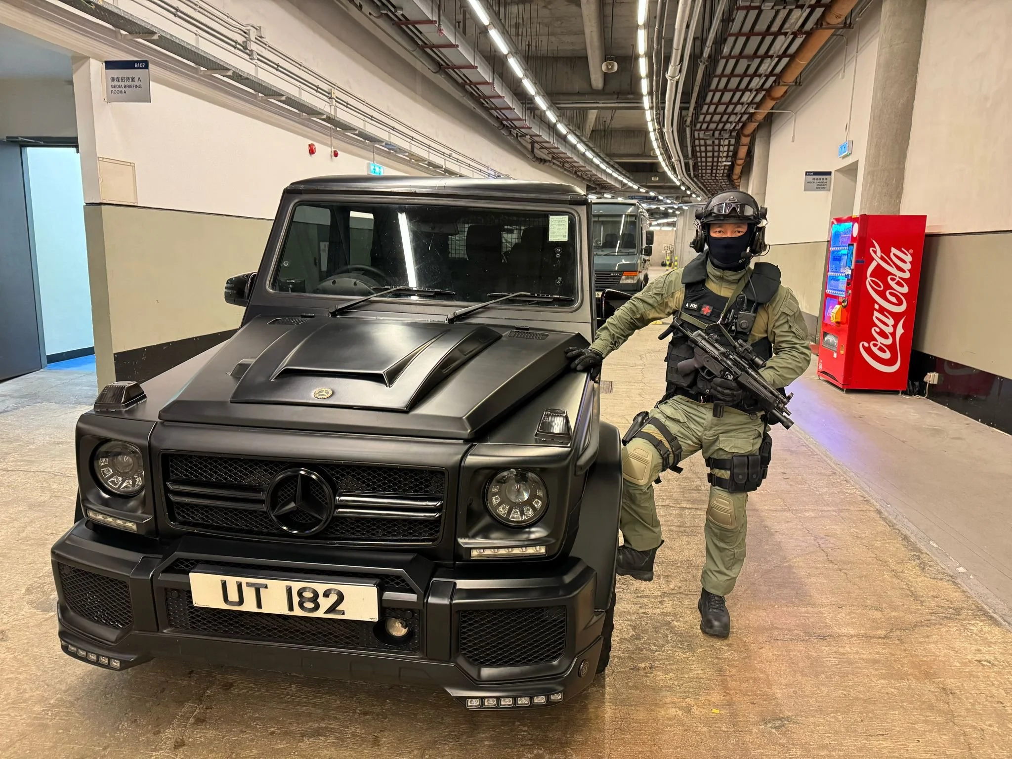 A tactical police officer in tactical gear, holding a rifle, leaning against a black Mercedes G-Class vehicle inside a parking garage, with a Coca-Cola vending machine in the background.