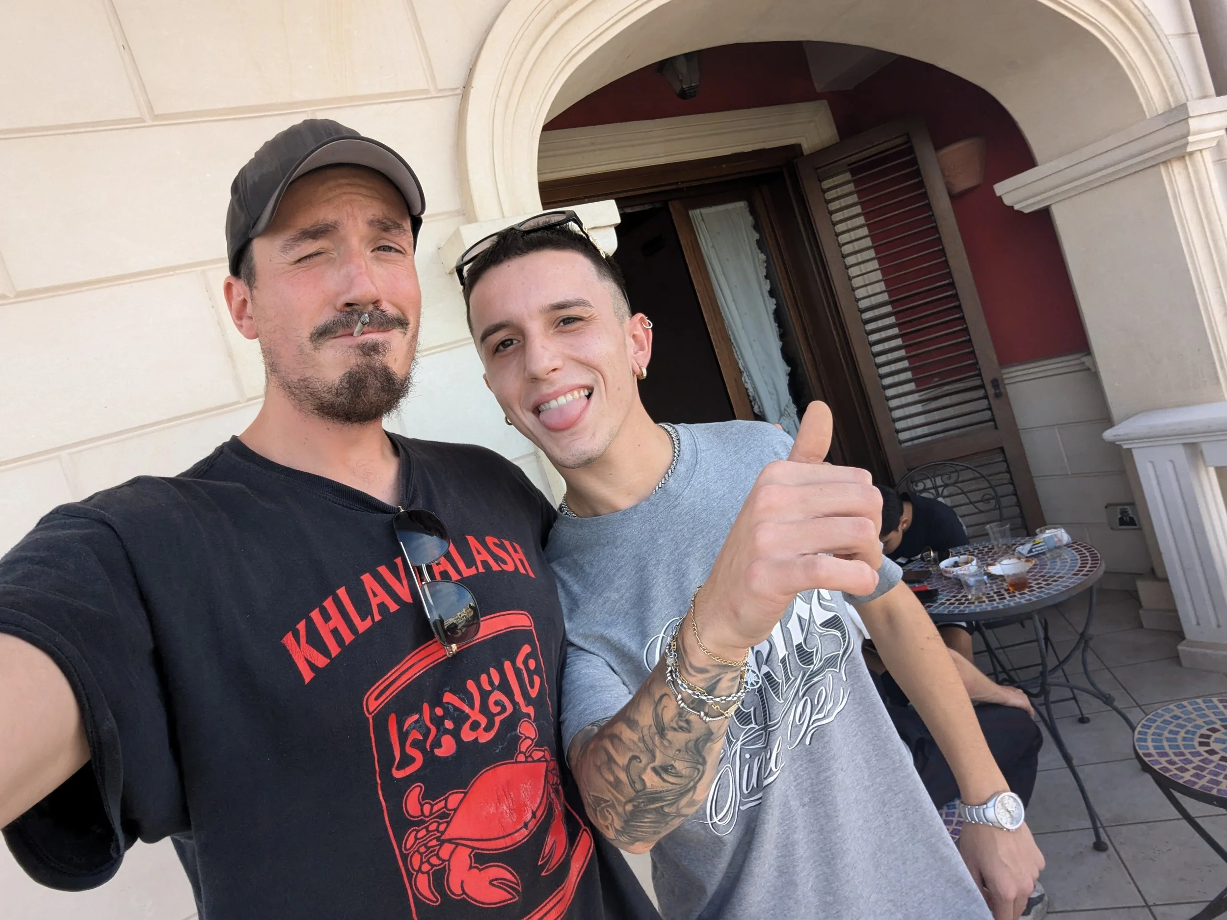 Two men taking a selfie outside on a patio. One is winking and smoking a cigarette, wearing a black T-shirt and baseball cap. The other is sticking out his tongue, wearing a gray T-shirt, bracelets, and jewelry. There is a table with drinks and a person sitting at the table in the background.