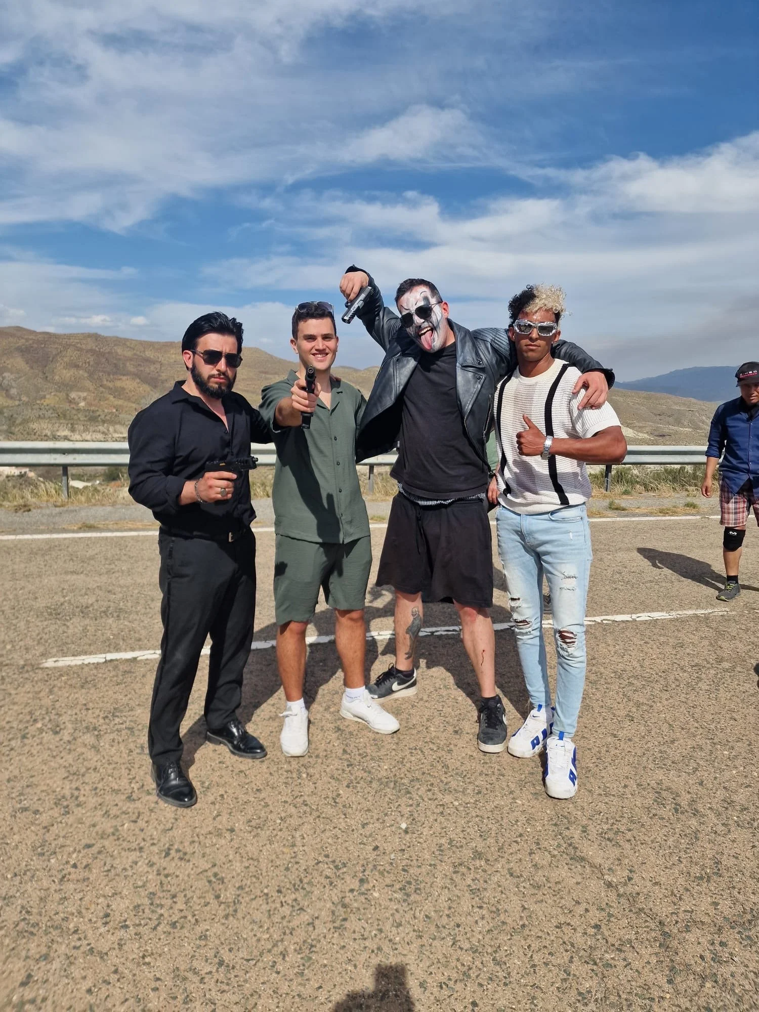 Group of five men standing on a road with mountains and blue sky in the background, some holding toy guns, one dressed as a gangster with face paint, wearing sunglasses and making a playful gesture.