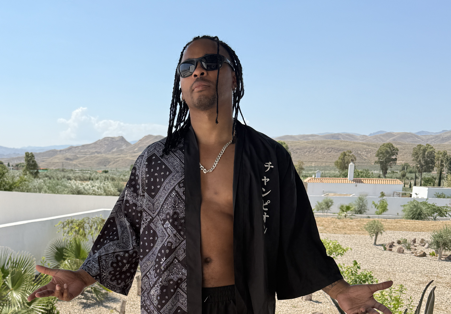 A man with braided hair and sunglasses stands with his arms outstretched in a desert landscape, wearing an open black and white patterned shirt and a silver chain necklace.