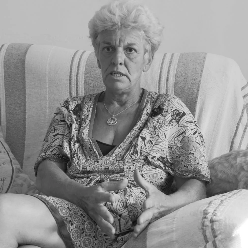 A black and white photo of an elderly woman with curly hair, sitting on a striped couch, making a hand gesture, wearing a patterned top and a pendant necklace.