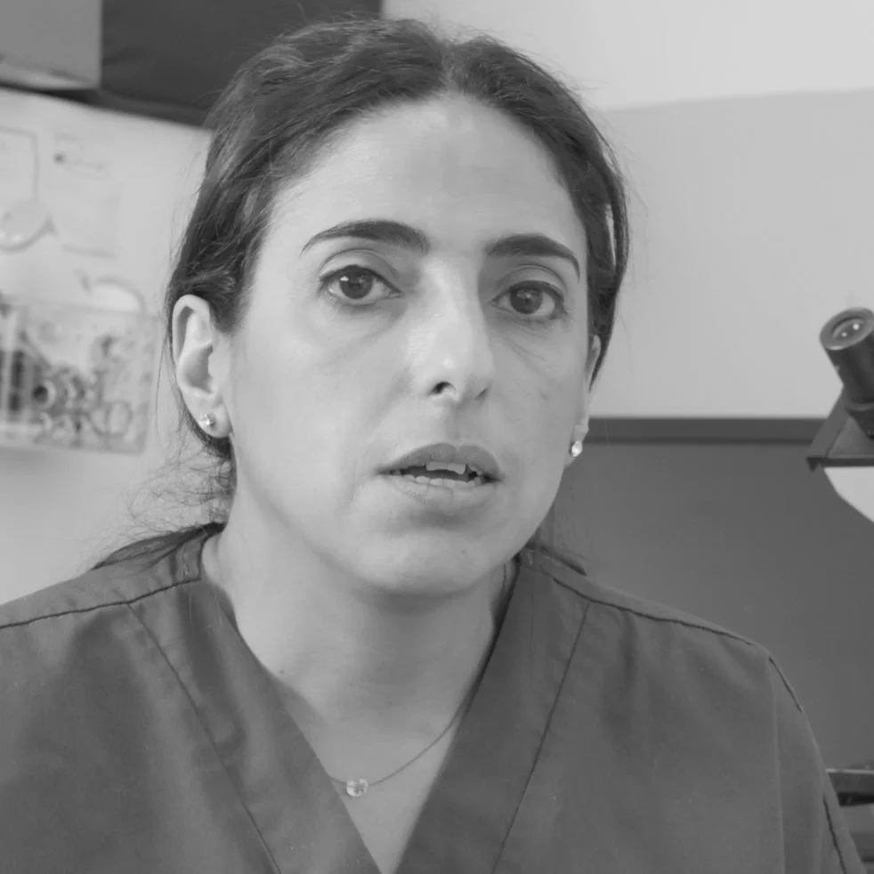 A woman with dark hair wearing medical scrubs, sitting in a clinical environment with a microscope and medical supplies in the background.