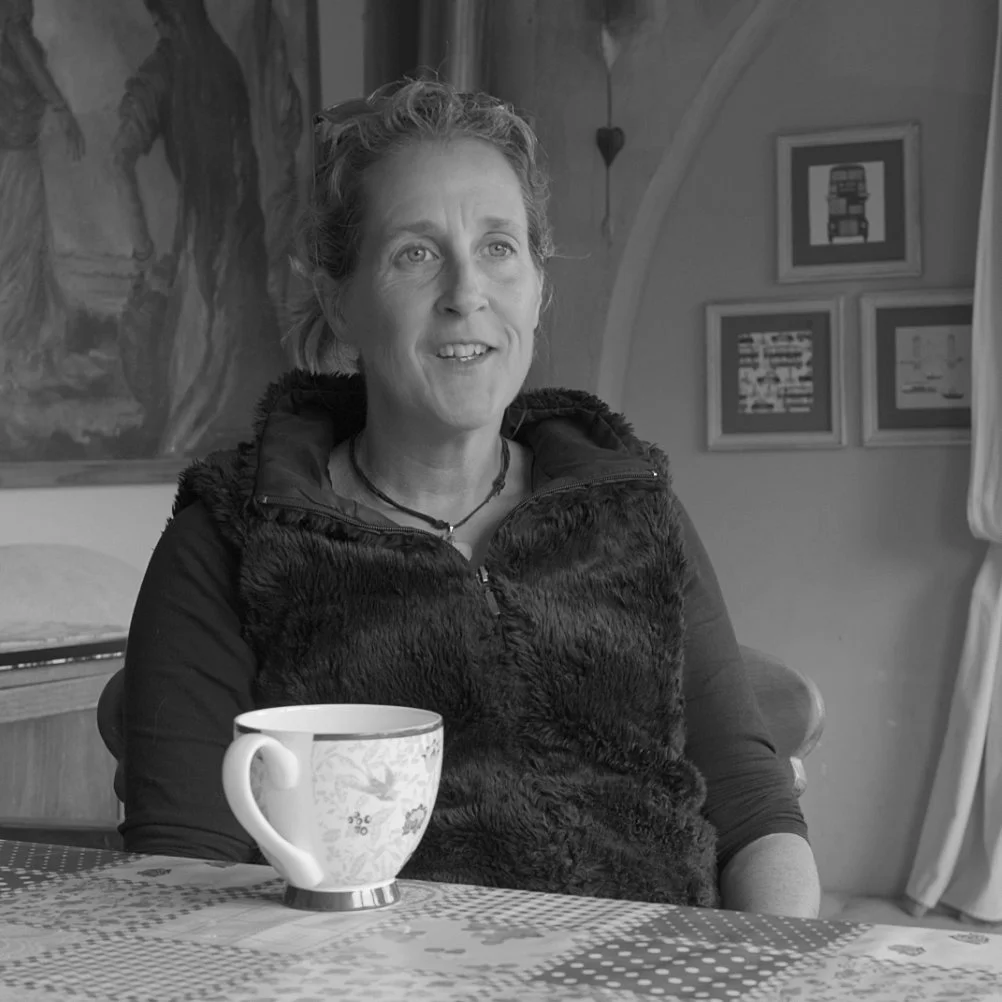 A woman sitting at a table with a coffee mug, smiling, in a room with framed pictures on the wall and a curtain.