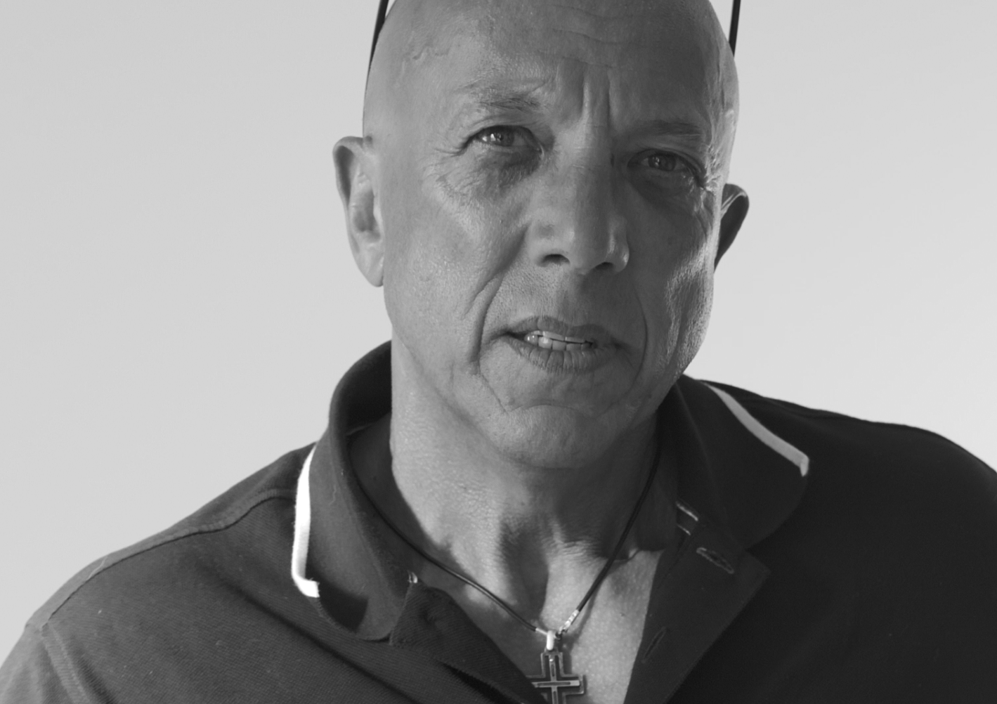 Black and white close-up of a middle-aged man with short hair, wearing a collared shirt, a necklace with a cross pendant, and earphones, against a plain background.