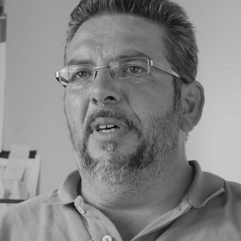 A middle-aged man with glasses, a beard, and mustache, wearing a collared shirt, speaking or explaining something.
