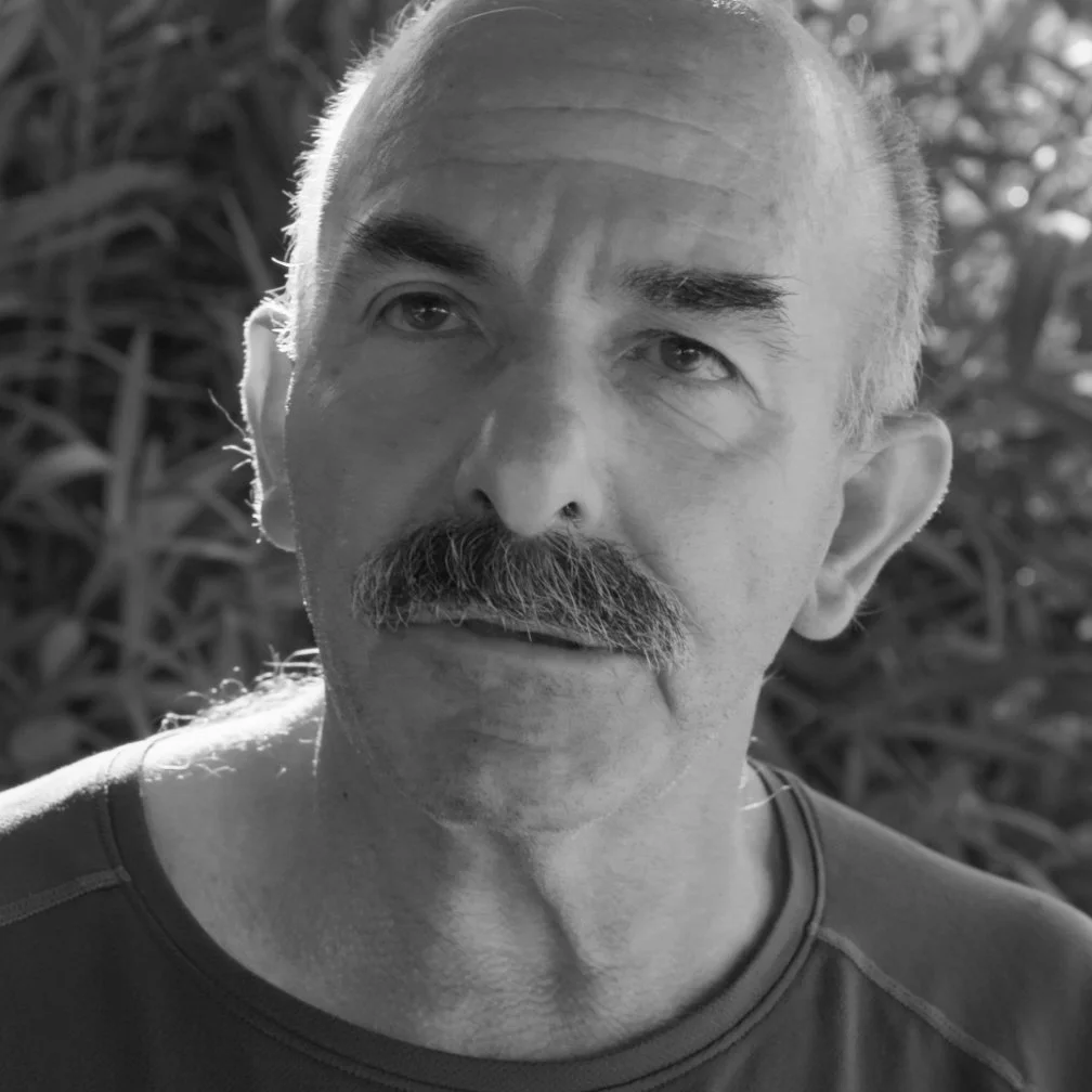 Black and white photo of a middle-aged man with a mustache and receding hairline, outdoors with trees and foliage in the background.