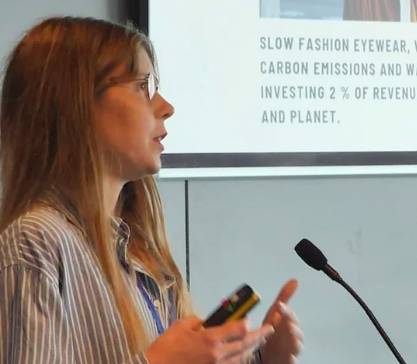 A woman with long red hair and glasses speaking at a presentation, holding a phone, with a slide about slow fashion eyewear and sustainability in the background.