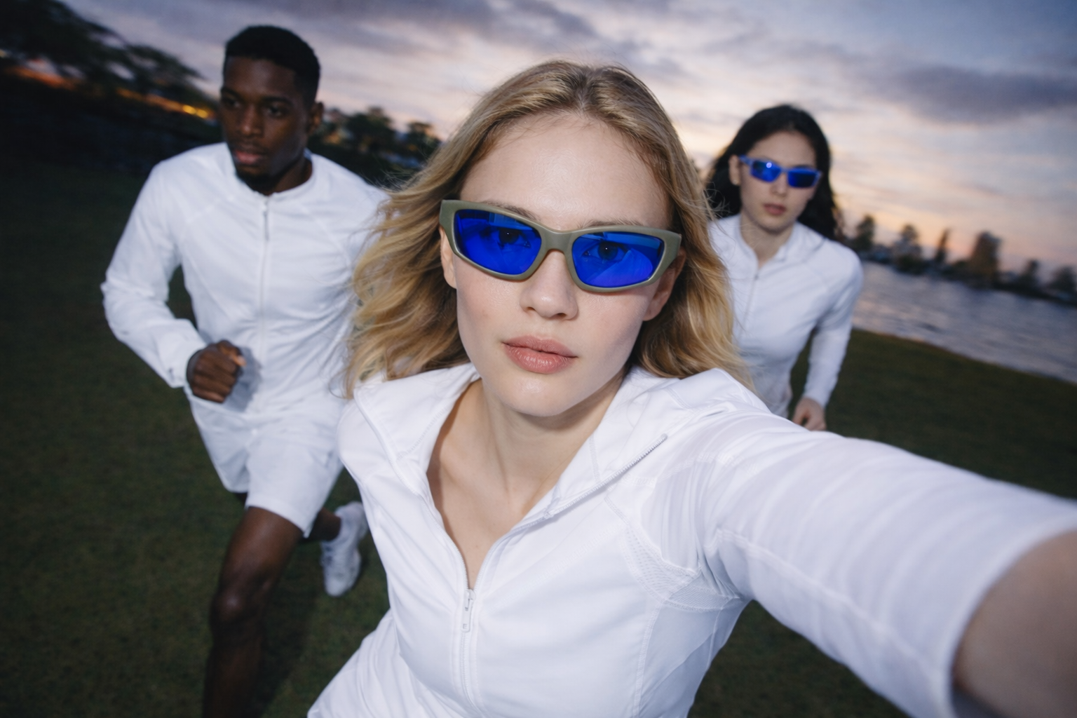 Three young women and a young man wearing white athletic outfits and blue sunglasses running outdoors near a body of water during sunset.