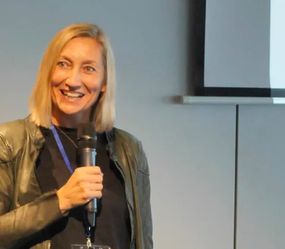 A woman with blonde hair smiling and holding a microphone, standing indoors next to a wall and window.
