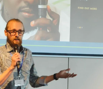 A man with glasses, beard, and a patterned shirt giving a presentation with a microphone. A projector screen behind him displays a close-up of a person holding a microphone and some text.