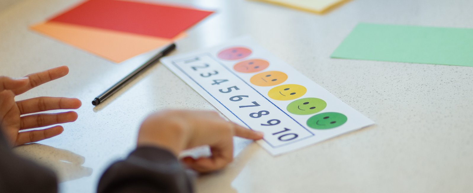 Ein Kind zeigt mit dem Finger auf eine Tischmatte mit einer Farbskala von roten bis grünen Smiley-Gesichtern und Zahlen von 1 bis 10, umgeben von Farbpapieren.