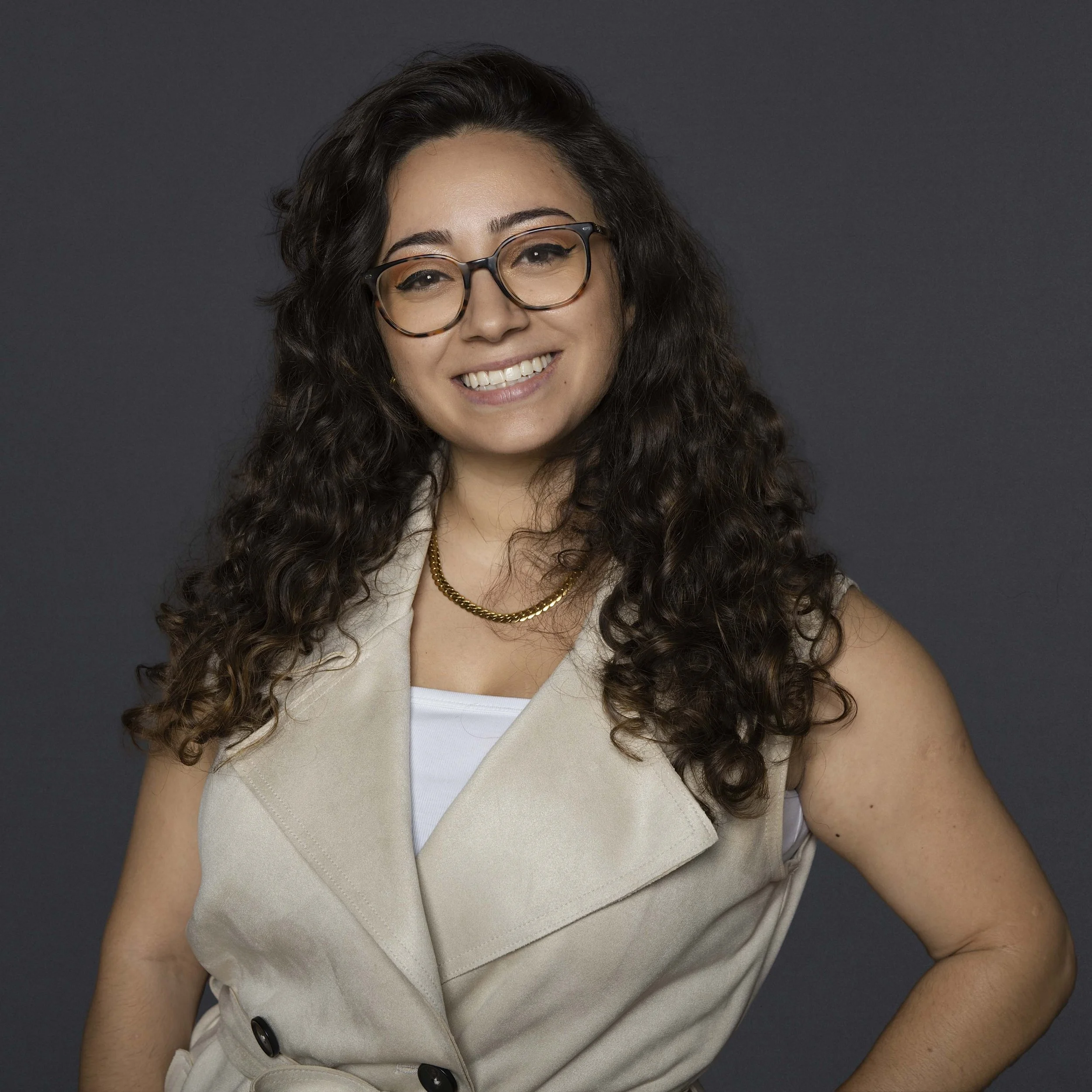 Une femme souriante portant des lunettes, un vêtement beige, une chaîne en or, et un t-shirt blanc, pose contre un fond noir.