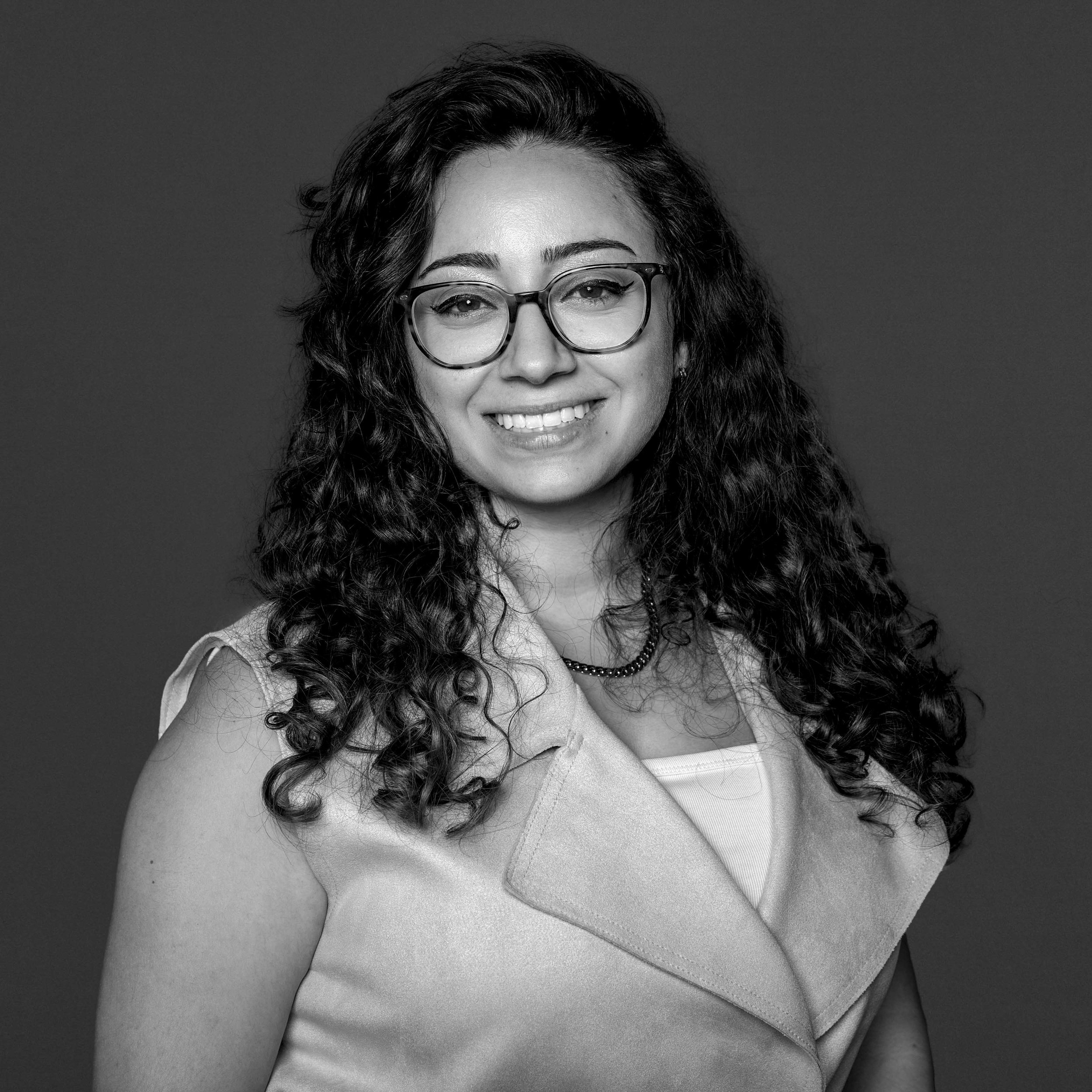 Portrait en noir et blanc d'une femme souriante avec des cheveux bouclés portant des lunettes, un col sans manches, et un collier