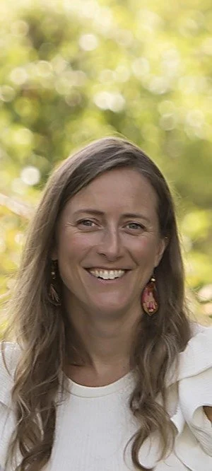 A woman with long brown hair smiling outdoors with a blurred green background.
