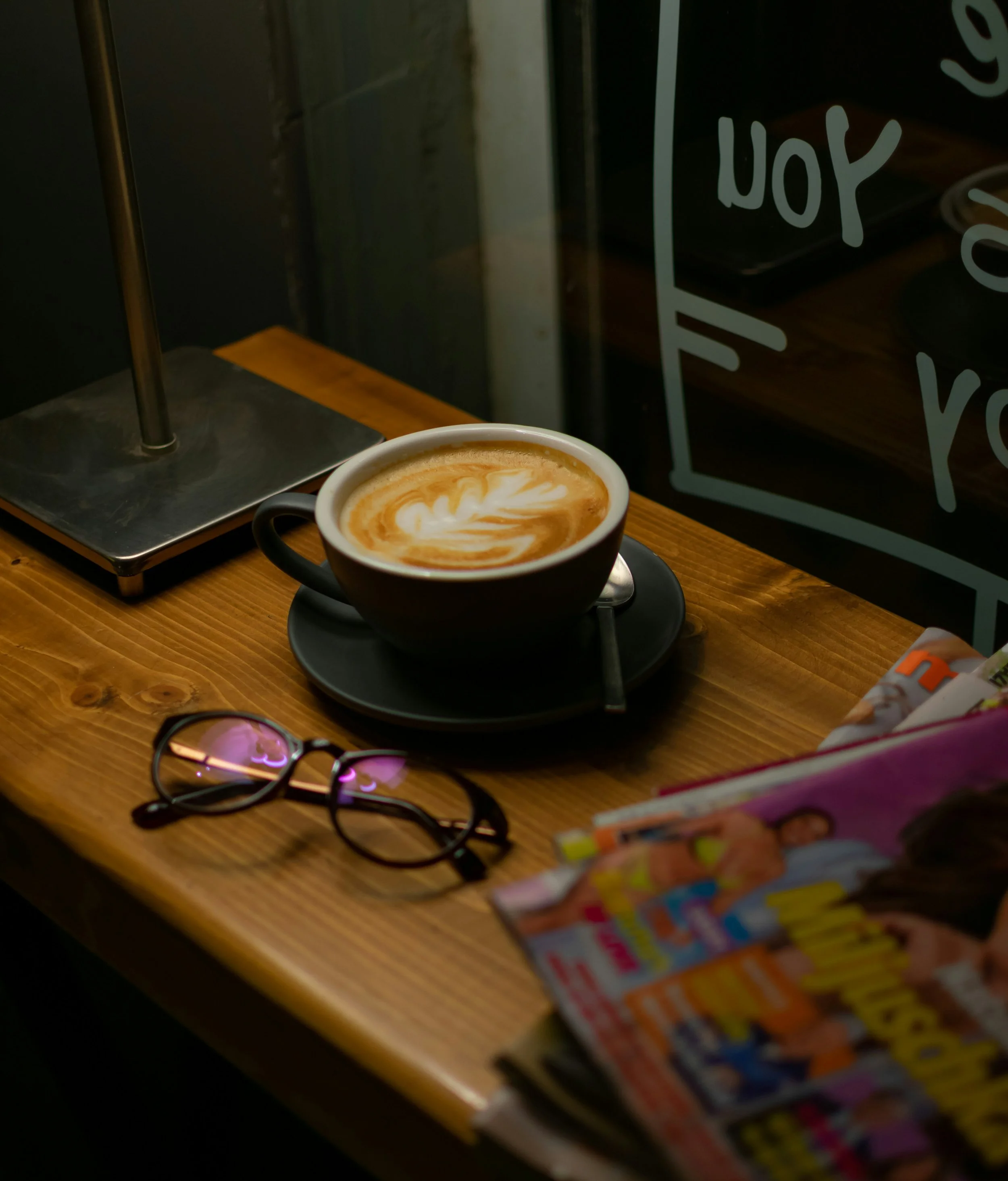 A cup of coffee with latte art on a wooden table near a window with magazine and a pair of glasses.