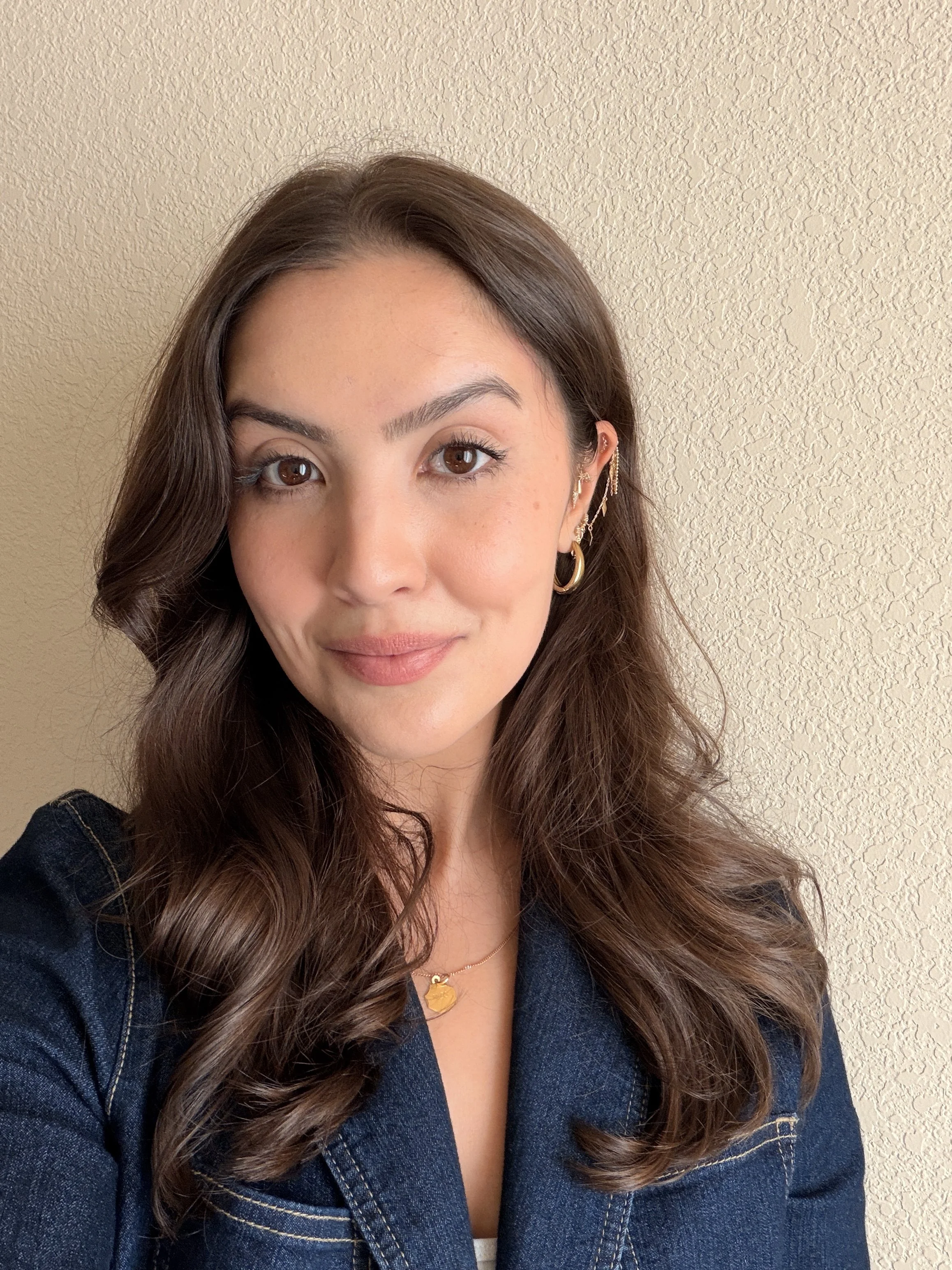 A young woman with wavy brown hair and brown eyes smiling at the camera, wearing gold hoop earrings, layered necklaces, and a dark blue blazer, standing in front of a beige textured wall.