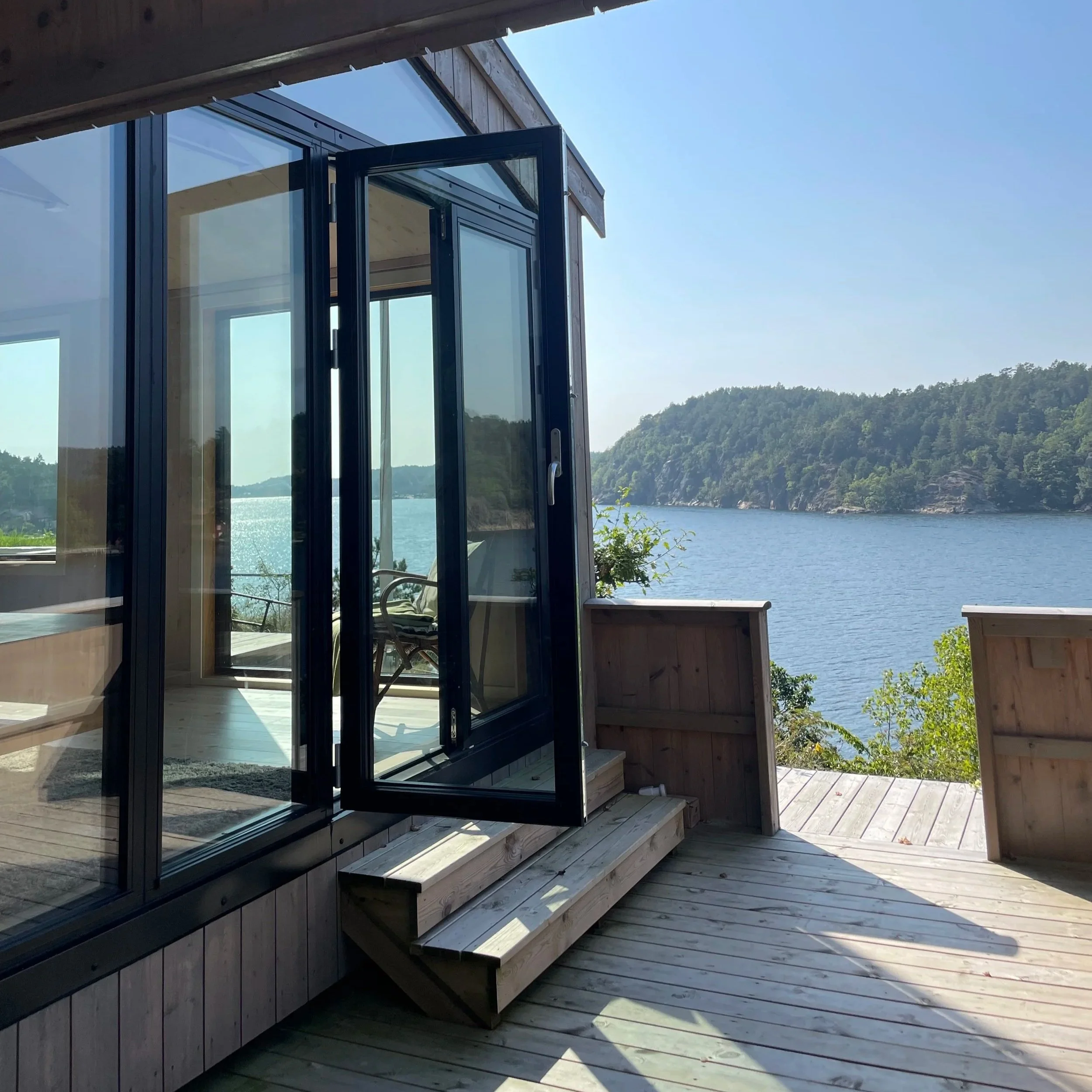 Utendørs terrasse med utsikt over vann og grønne fjell, moderne glassdør og trevegg.
