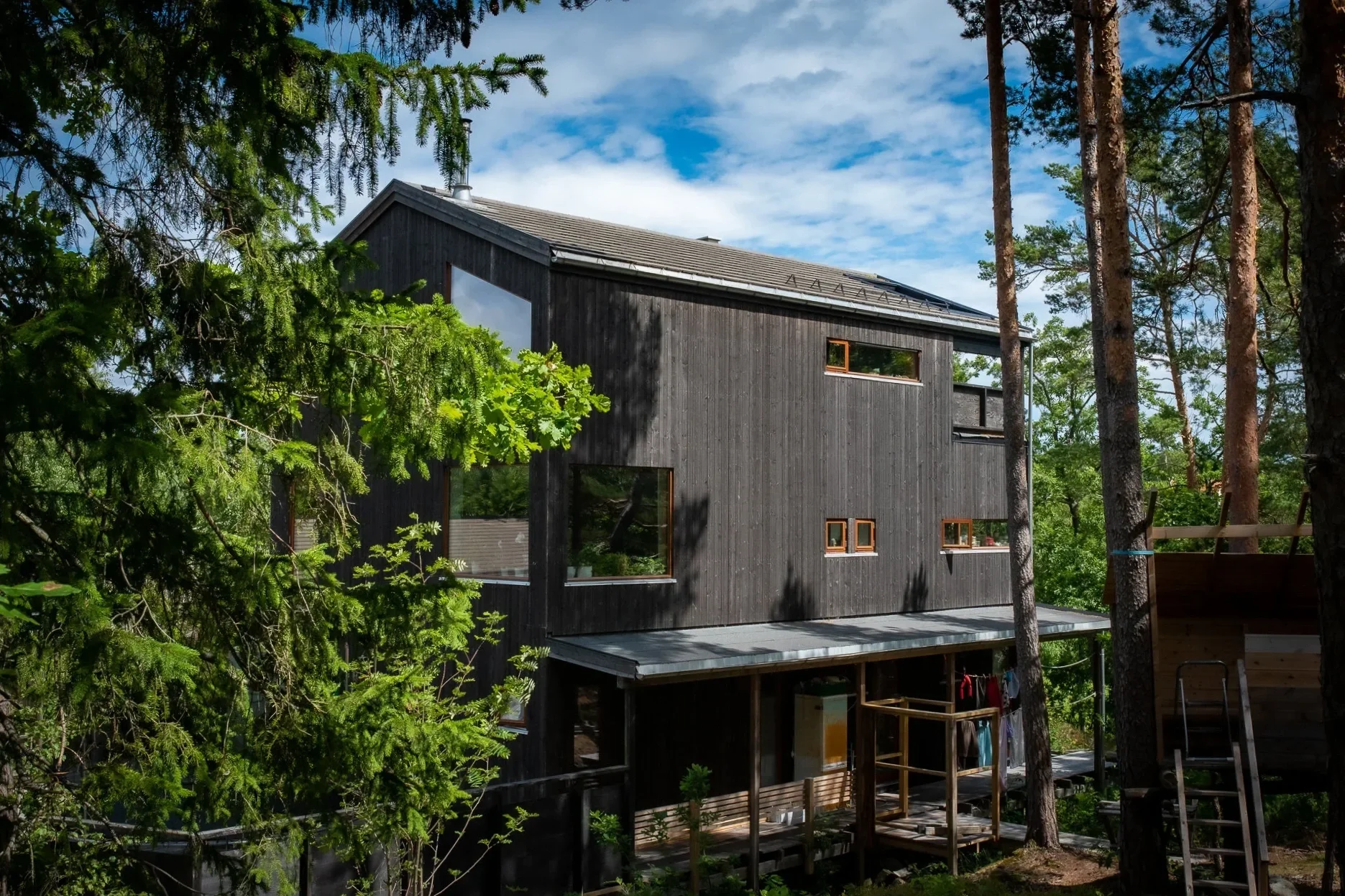 Et moderne trehus i skogen, omgitt av grønne trær under en blå himmel med noen skyer.