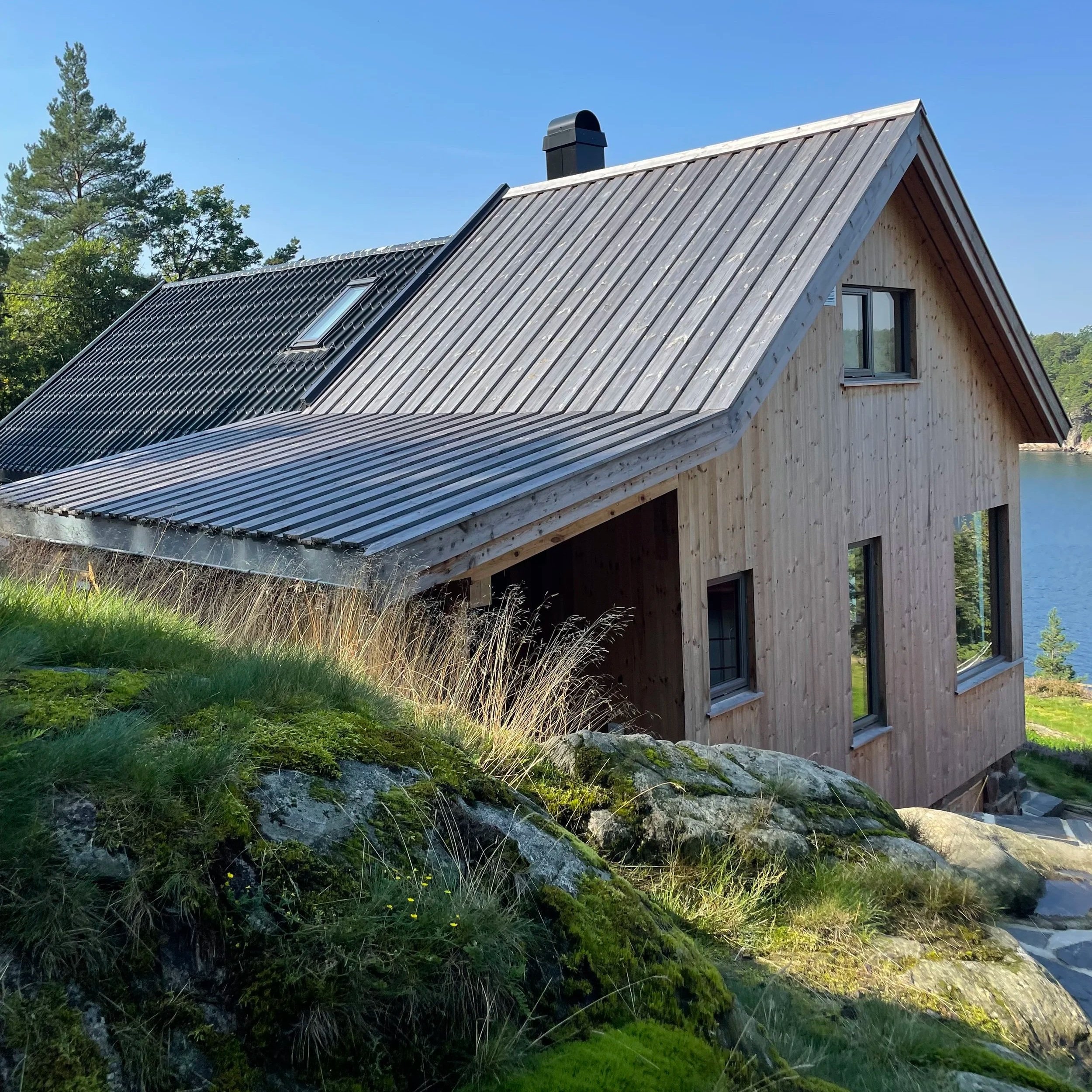 Nærbilde av et moderne bolig med treutvendig kledning, store vinduer og brunt tak. Husets omgivelser inkluderer grønne planter, gress og steiner, med klare blå himmel i bakgrunnen.