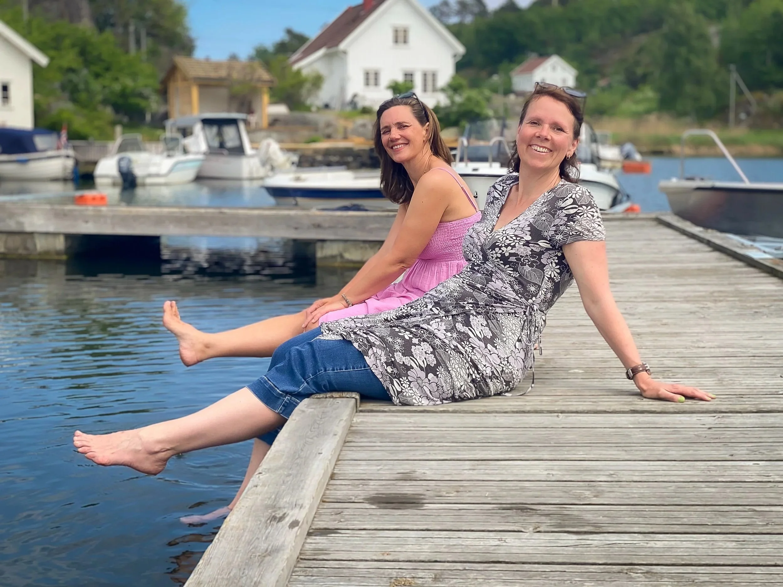 To arkitekter sitter på en brygge ved vannet med båter i bakgrunnen, smilende og nyter en sommertid ved sjøen.