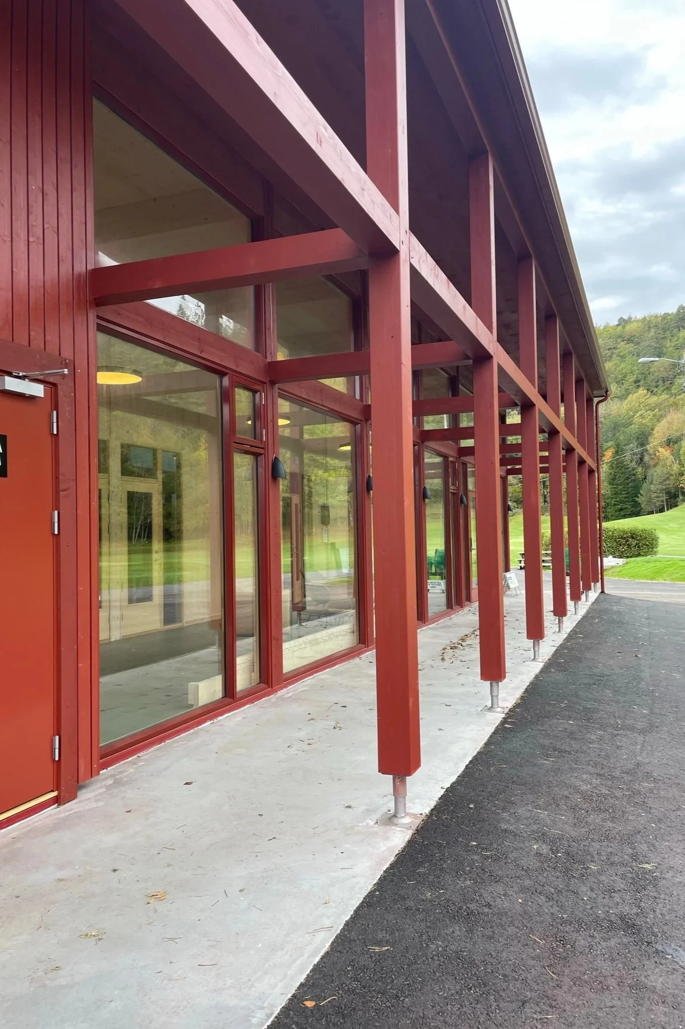En moderne offentlig bygning med store glassvinduer og røde trebjelker. Uteområdet har en asfaltvei og en betongflate, omgitt av grønt landskap og trær i bakgrunnen.