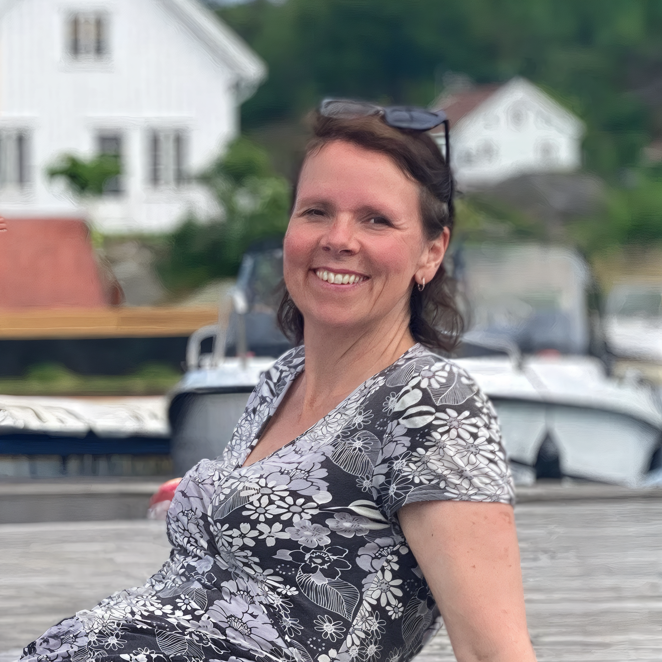 En kvinne (Ingeborg Marcussen) smiler utendørs foran hus og trær, iført svart og hvitt blomsterkjole og har en hundehalskjede. Også en datamaskin er synlig i bakgrunnen.