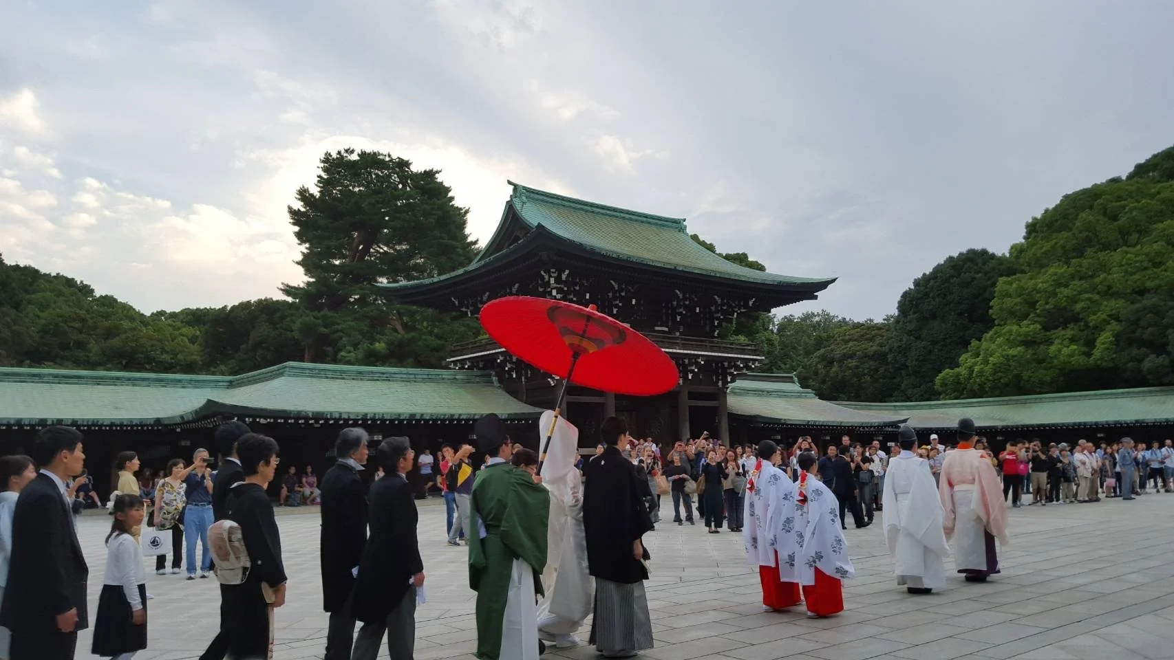 Cérémonie traditionnelle japonaise avec moines portant des kimonos dans un lieu historique entouré de personnes, avec un grand bâtiment en arrière-plan et des arbres verts.
