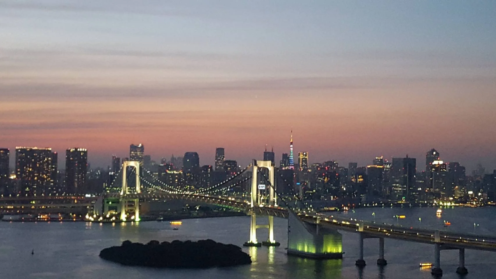 Vue de la ville de Tokyo au crépuscule, avec le Rainbow Bridge illuminé en premier plan et la tour de Tokyo illuminée au loin.