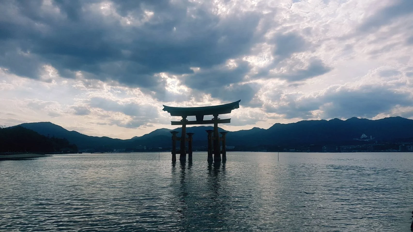 Une torii traditionnelle japonaise en bois, située dans l'eau, avec des montagnes en arrière-plan sous un ciel nuageux.