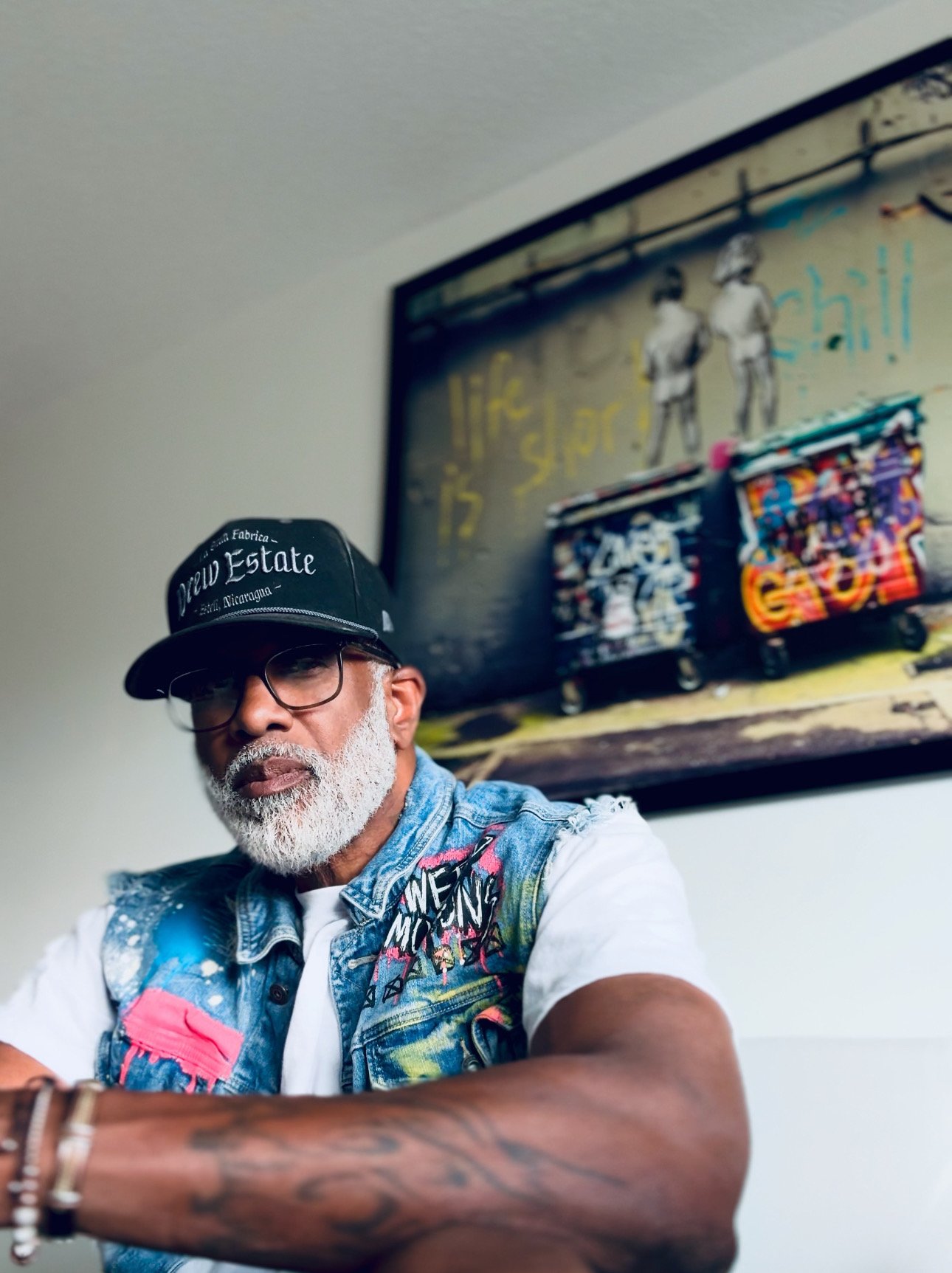 Man with a gray beard and glasses wearing a black cap, white shirt, and colorful denim vest sitting indoors in front of a colorful painting on a white wall.